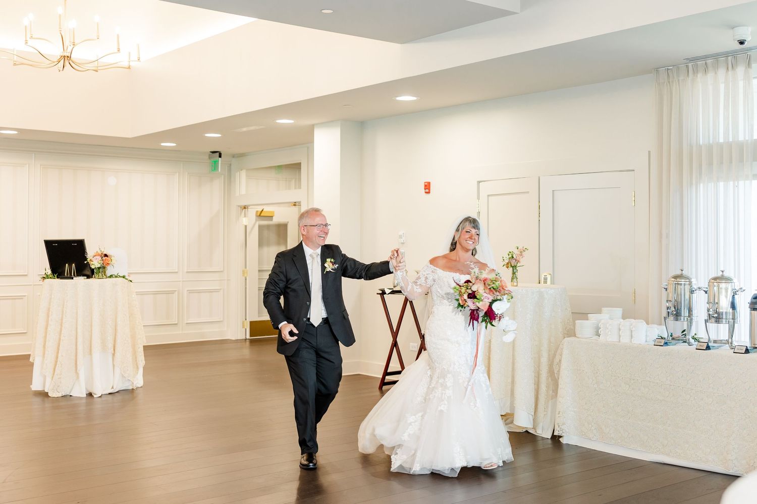 Bride in a lace wedding gown holding a bouquet walks hand in hand with her groom into a bright, elegant reception hall.