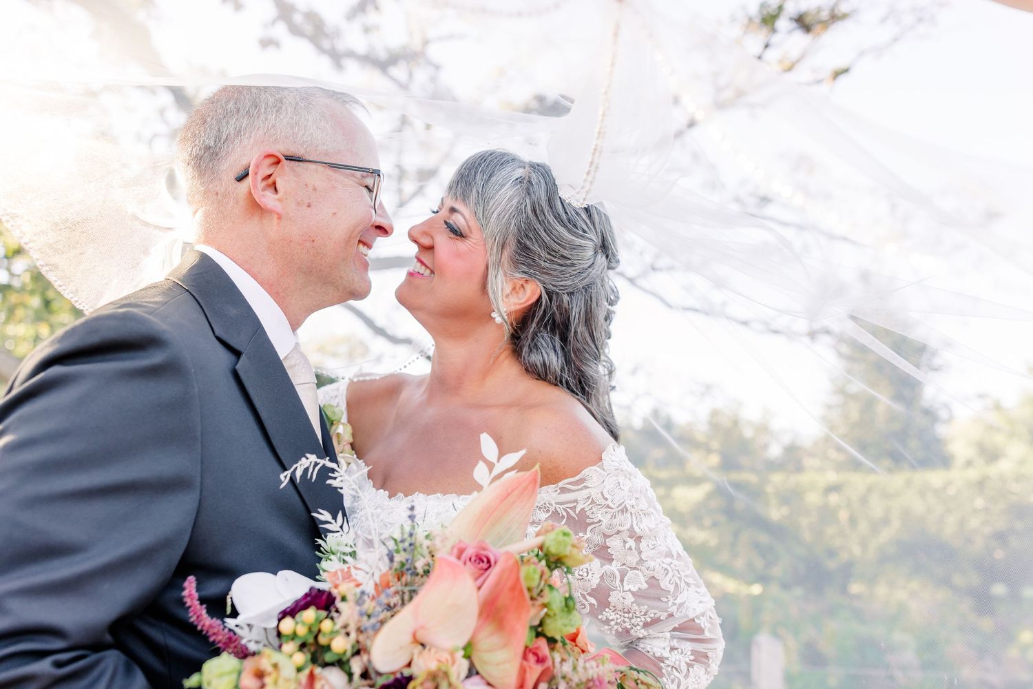 A groom in a gray suit and a bride in a lace gown smile at each other under a veil holding a colorful wedding bouquet.