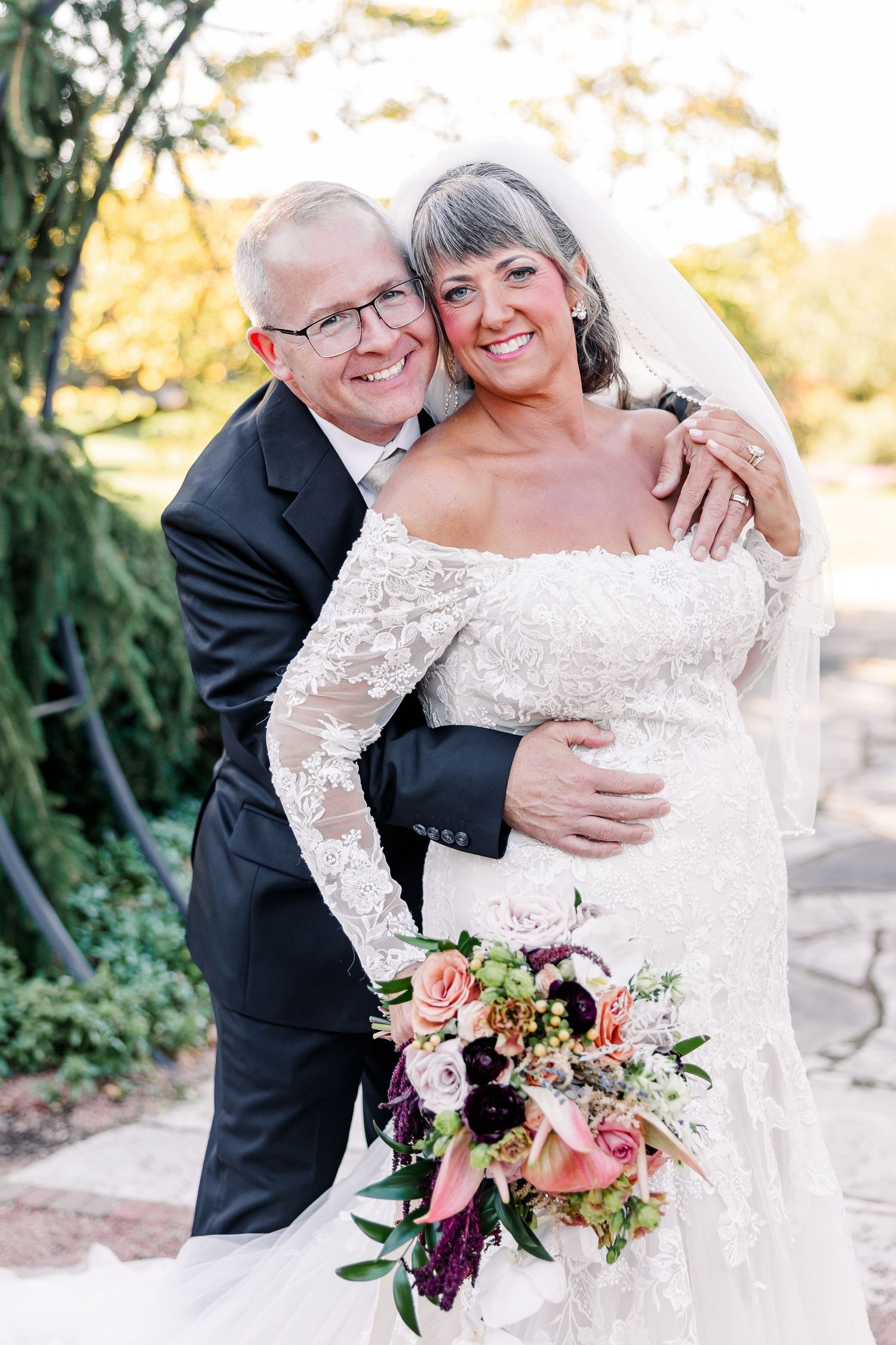 Smiling bride in a lace off-the-shoulder wedding gown and veil holding a colorful bouquet as her groom in glasses embraces her outdoors.