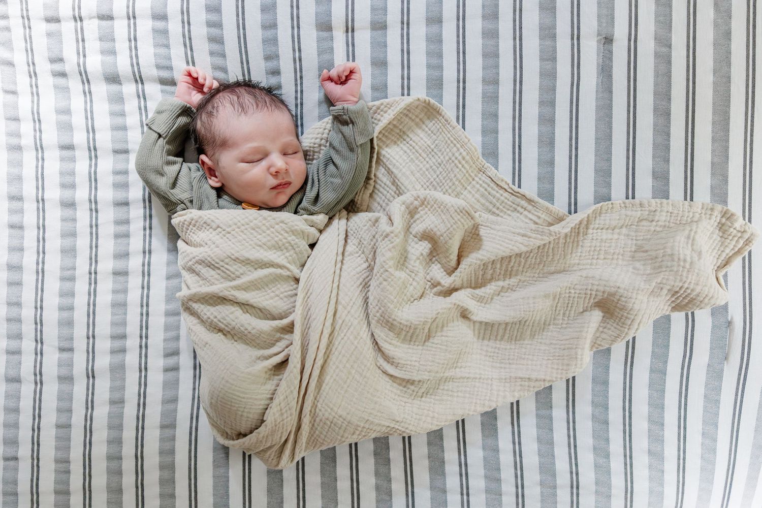 Newborn baby sleeping peacefully on striped blanket, arms raised, wrapped in soft cream muslin swaddle cloth.