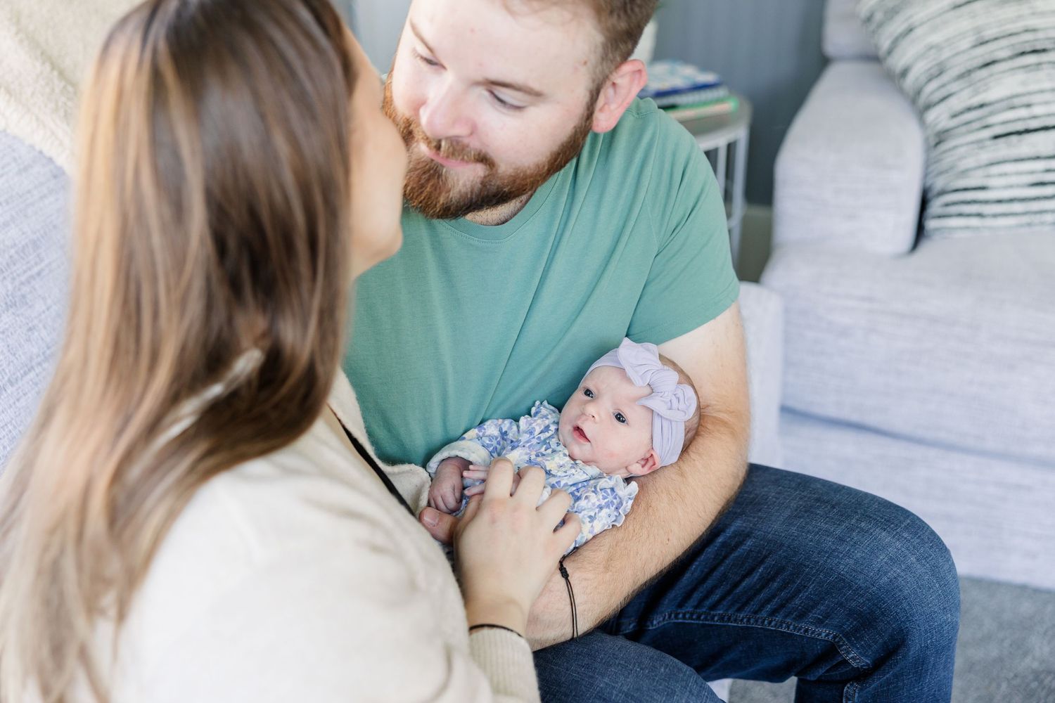 New parents smiling at their newborn baby girl in a floral outfit and pink headband during an in-home newborn session.