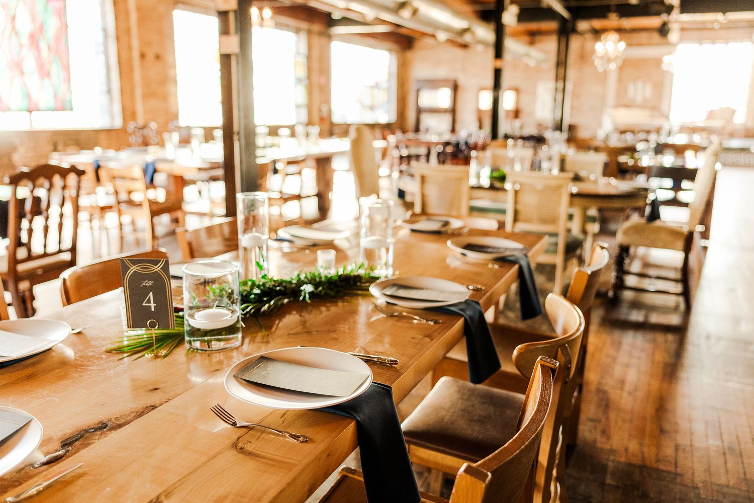 Long wooden dining tables set with white plates and greenery in sunlit restaurant.