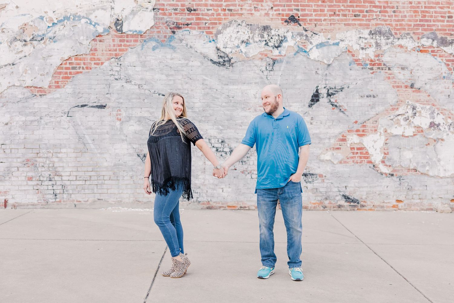 A couple holds hands while posing playfully in front of a weathered brick wall.