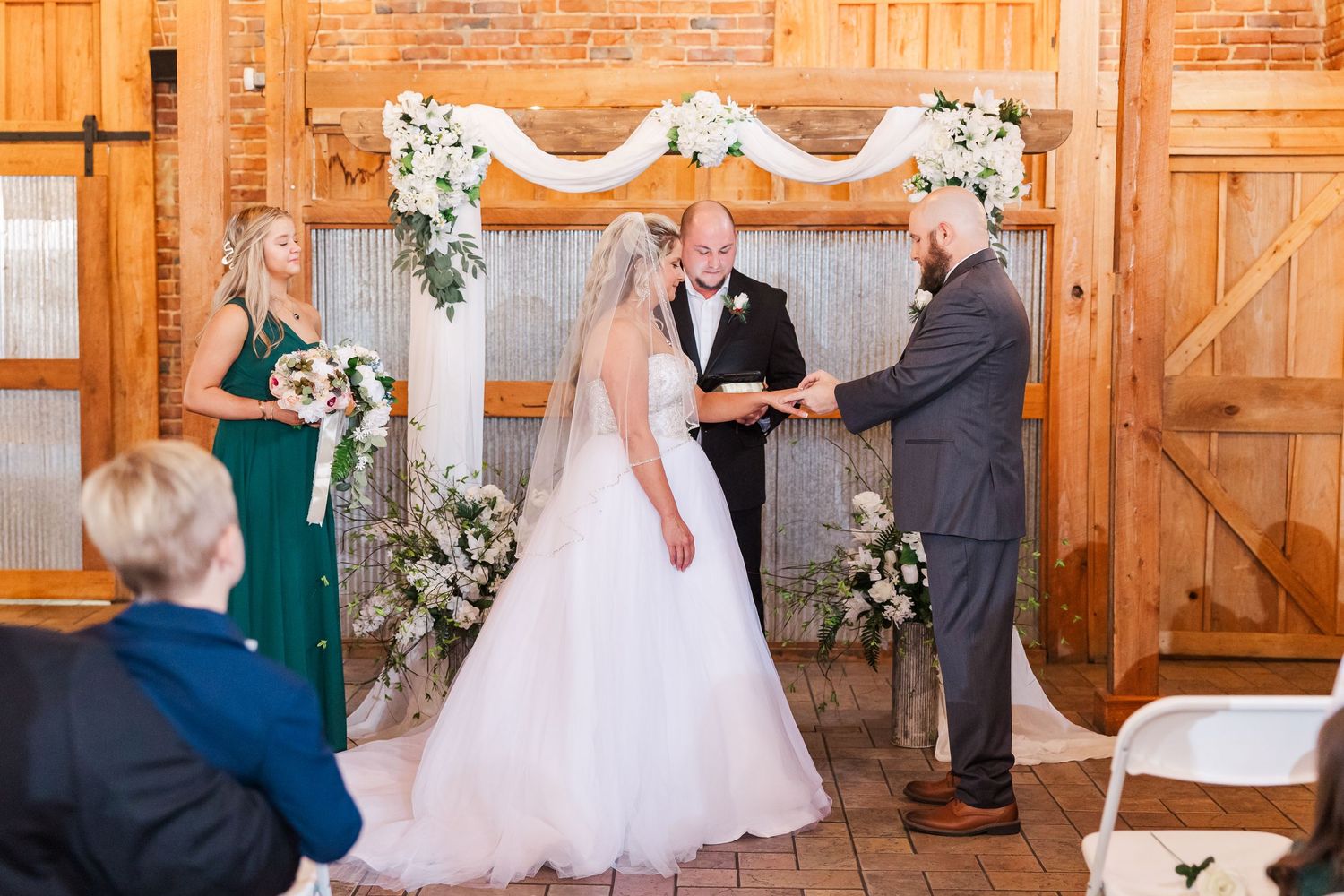 An intimate wedding ceremony takes place under a white floral arch in a rustic barn setting.