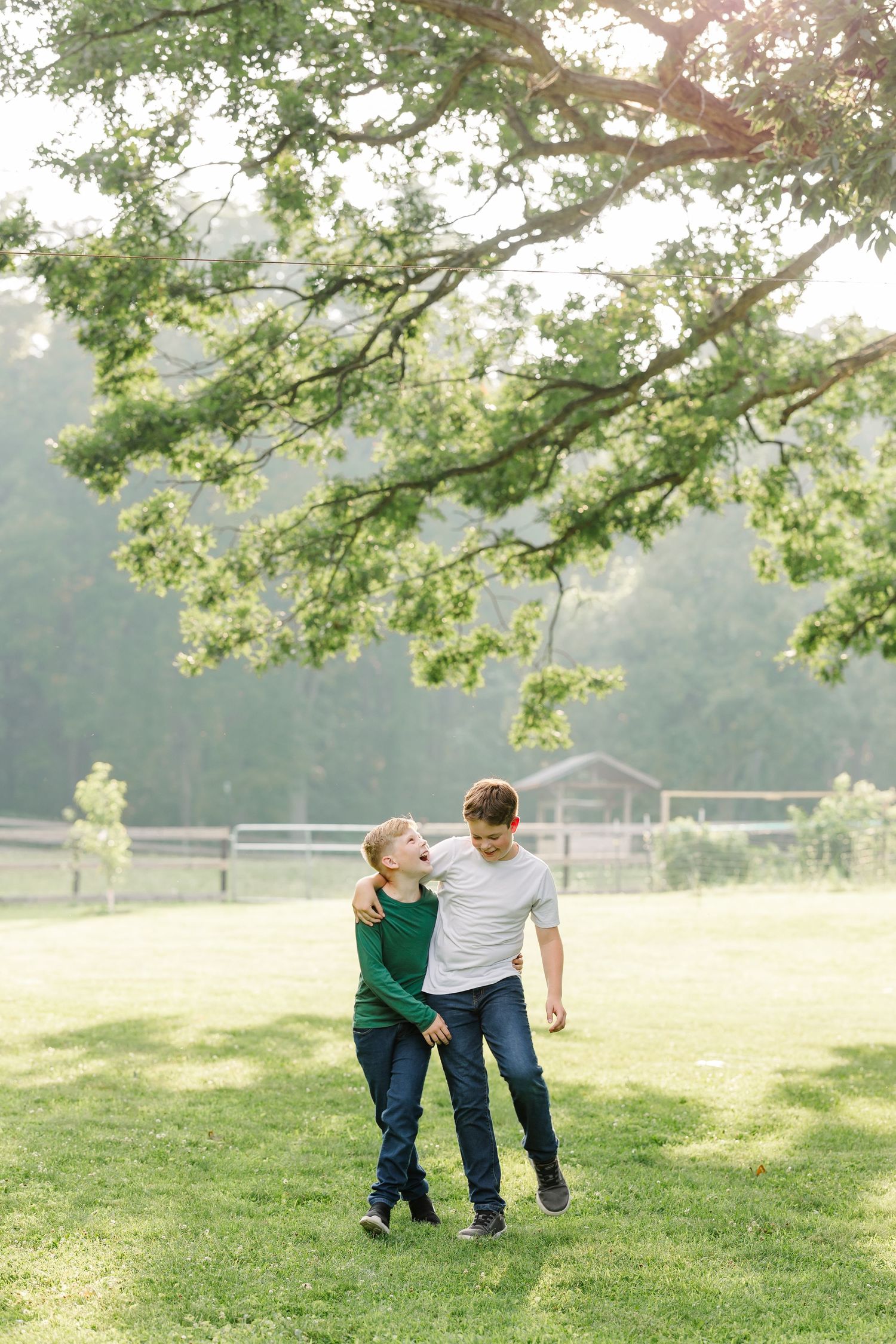 Two boys laughing and hugging under a large oak tree in a sunny green park.