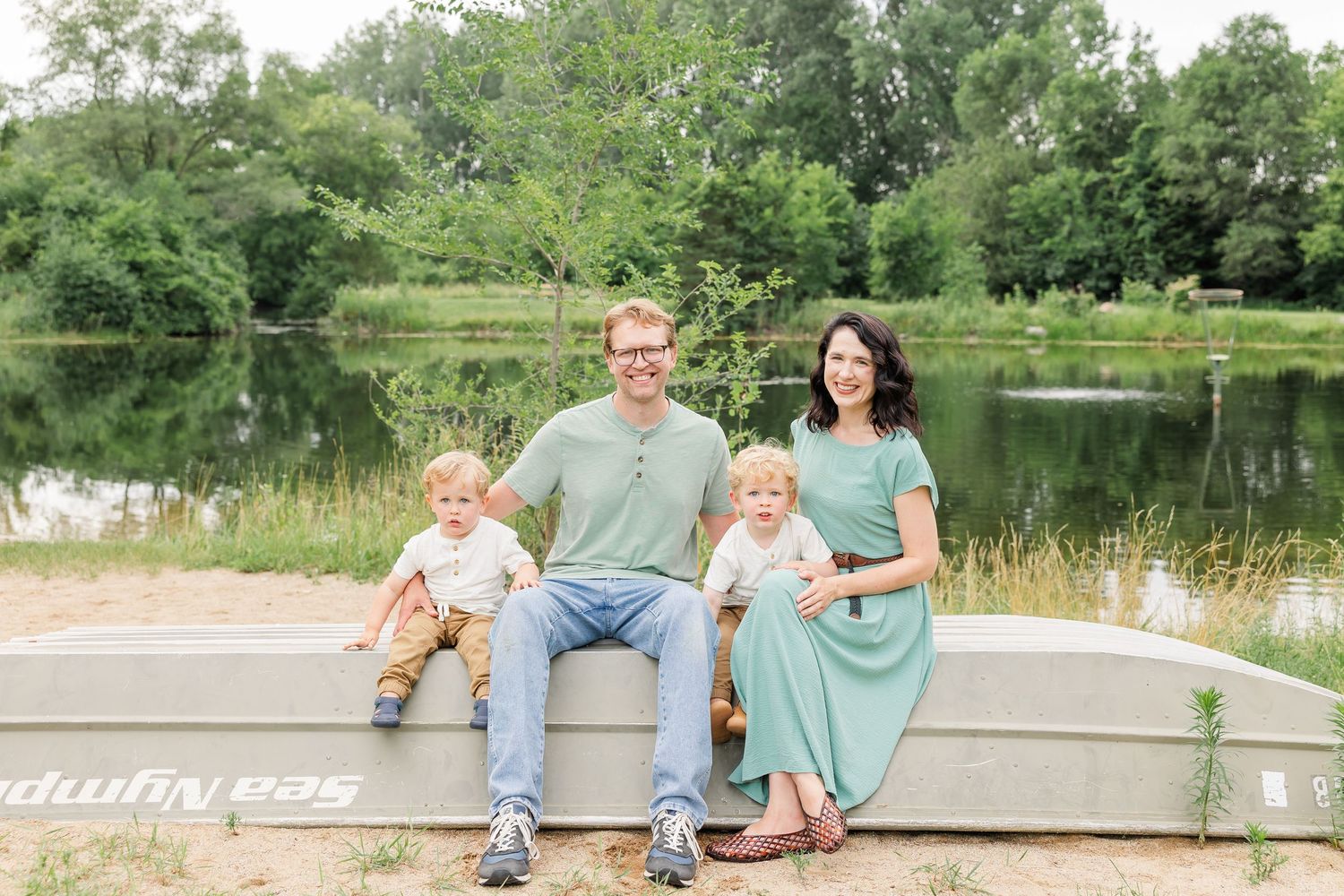 A smiling family of four sits on a boat by a calm reflective pond surrounded by green trees.