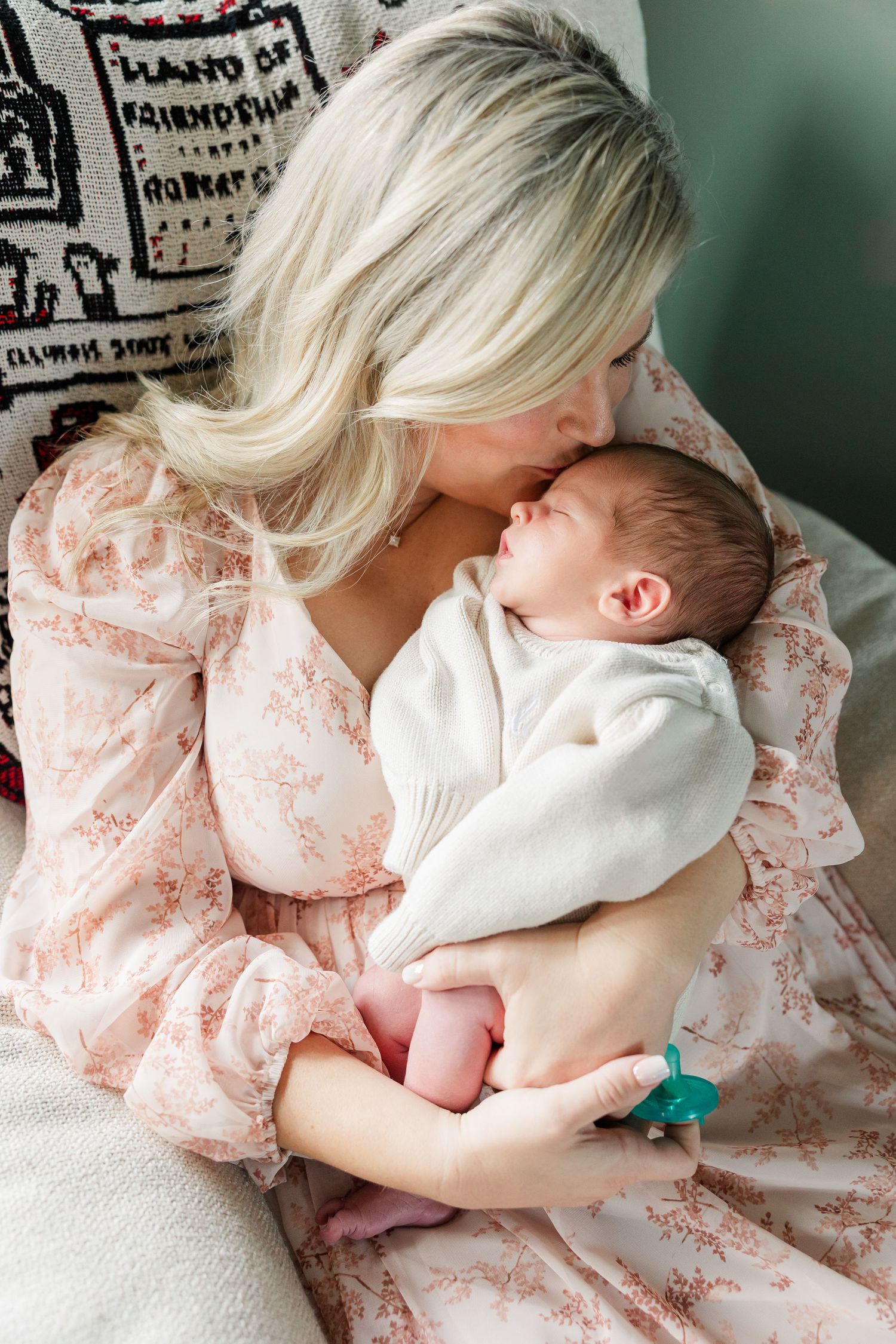 Blonde mother in floral dress kissing sleeping newborn baby wrapped in white swaddle.