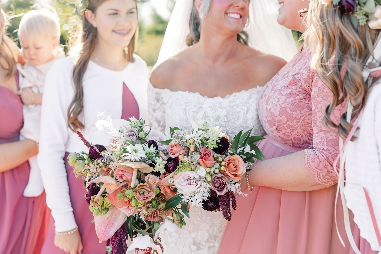 Bridesmaids in blush pink dresses stand beside a bride in white lace, with a lush bouquet of peach roses and greenery in front.