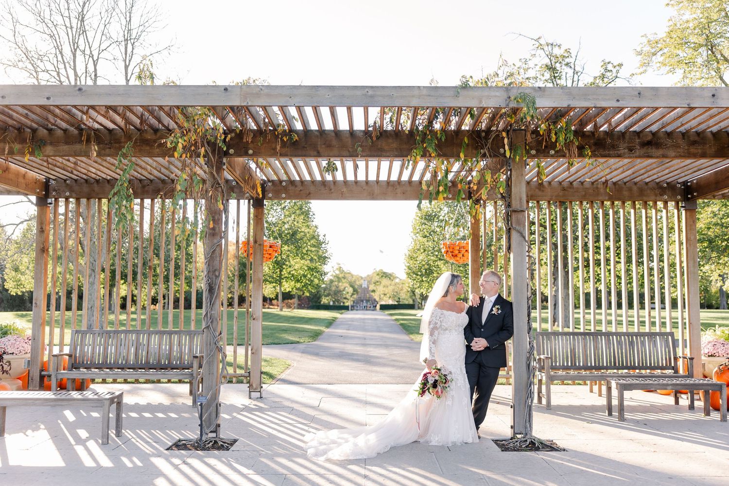 Bride and groom embrace beneath a wooden pergola with hanging greenery and wooden benches on a sunlit outdoor wedding venue patio.