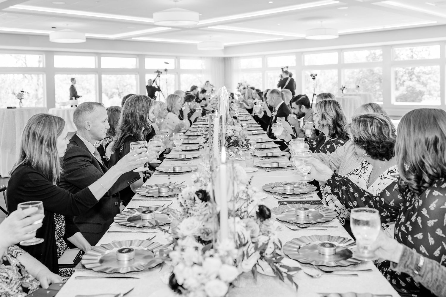 Black and white photo of wedding guests seated along a long reception table with floral centerpieces, place settings, and wine glasses.