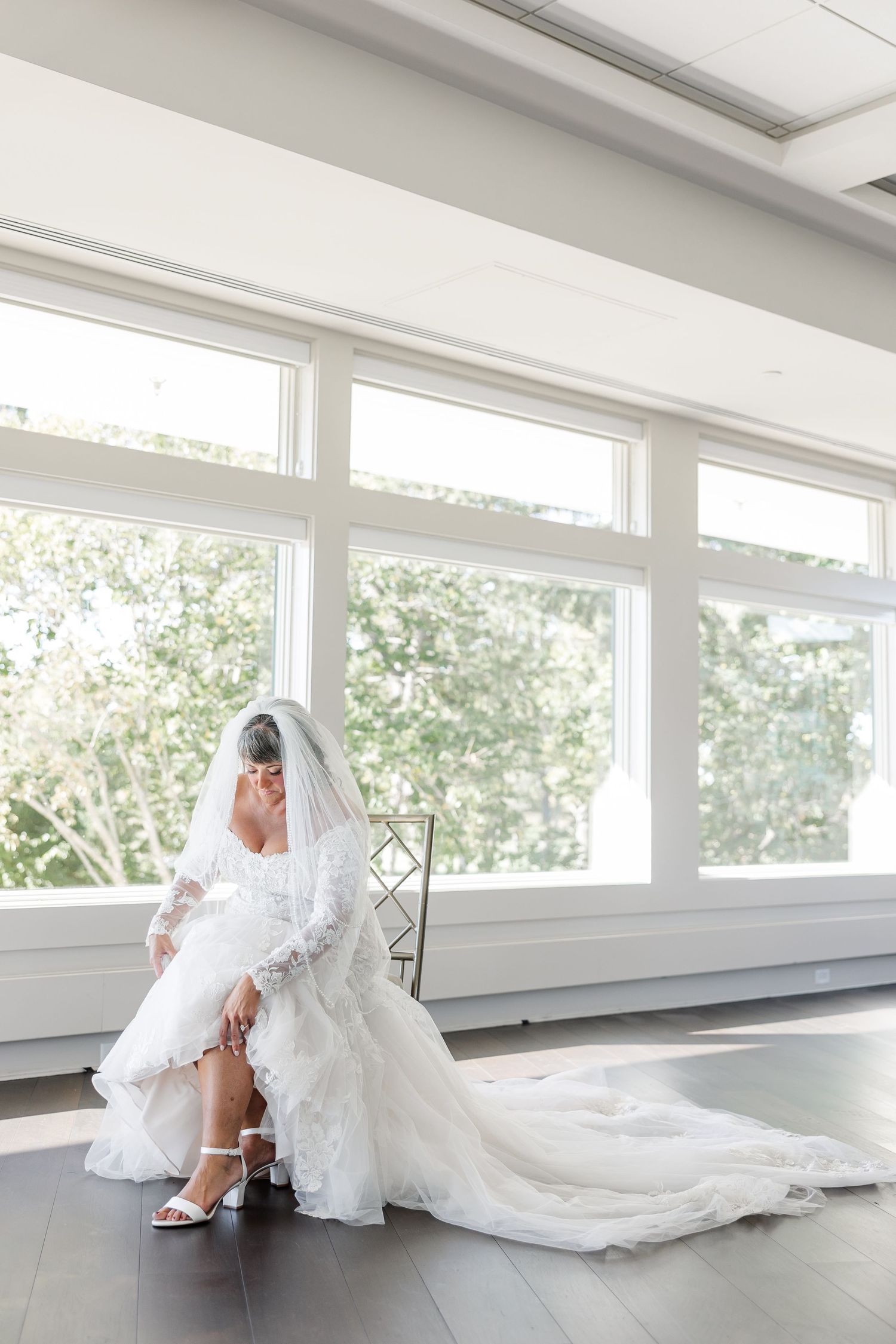 Bride in a lace long-sleeve wedding gown and veil sits adjusting her white strappy heel beside large sunlit windows.