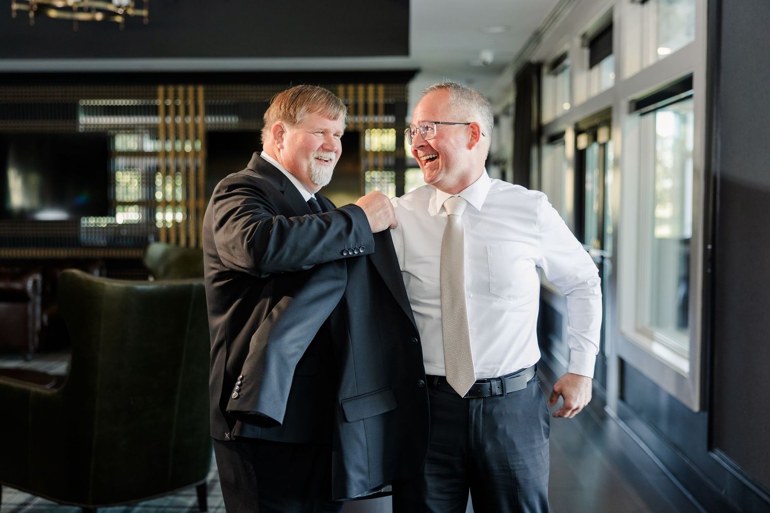 Two smiling men in formal attire share a warm moment in a modern, elegantly lit interior, one adjusting his dark suit jacket.