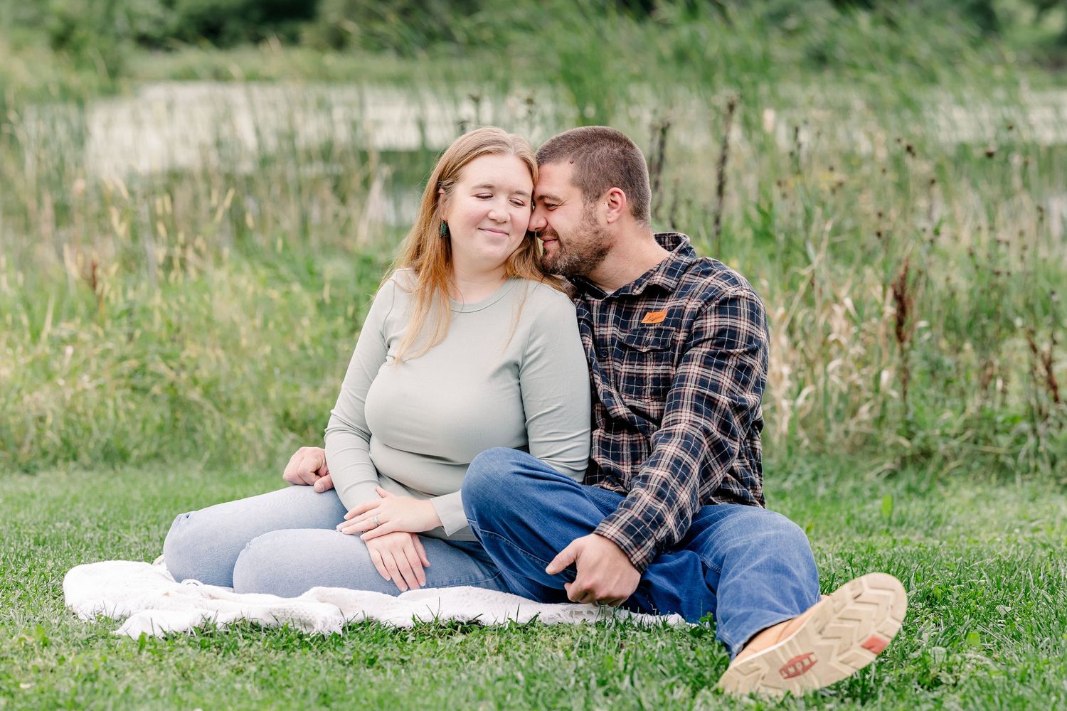 Couple sitting on a blanket in a grassy field, man kissing woman on cheek, outdoor engagement photo.