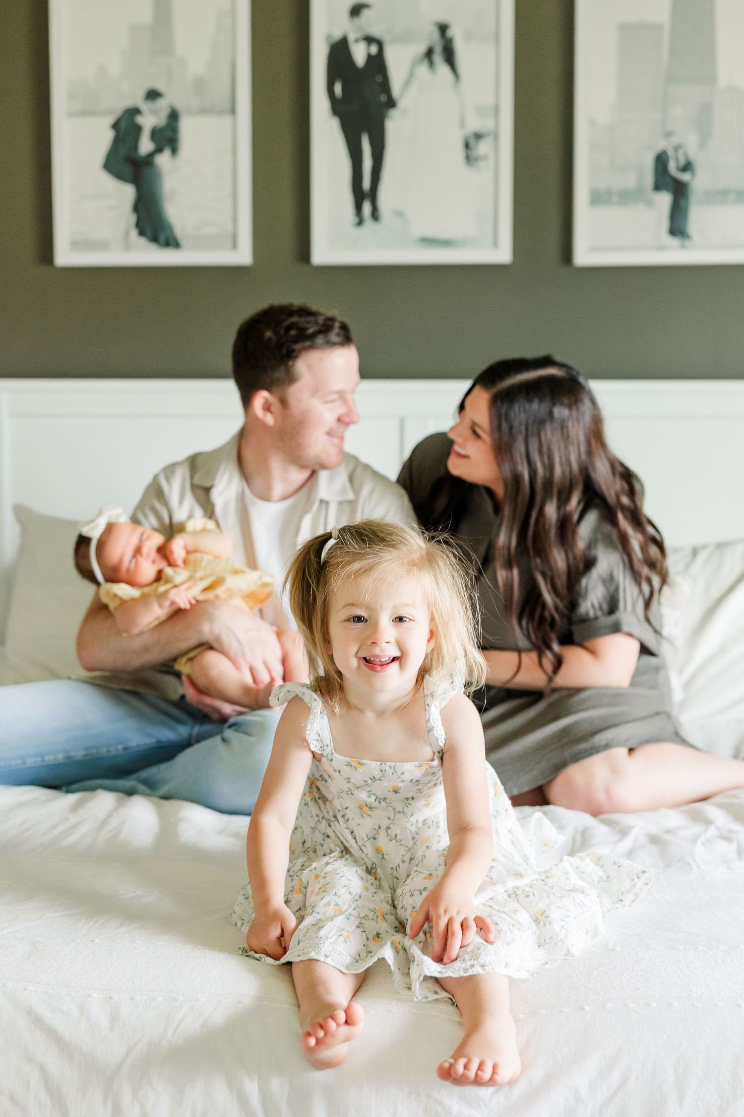 Big sister smiling on the bed while parents snuggle newborn baby girl during a lifestyle newborn session at home