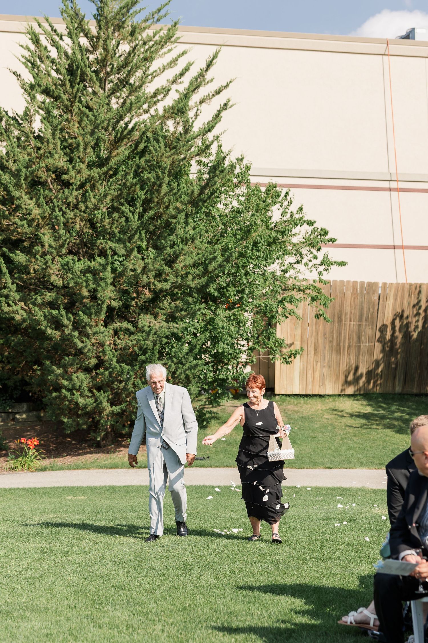 Elderly couple walks hand in hand across green lawn at outdoor wedding venue.