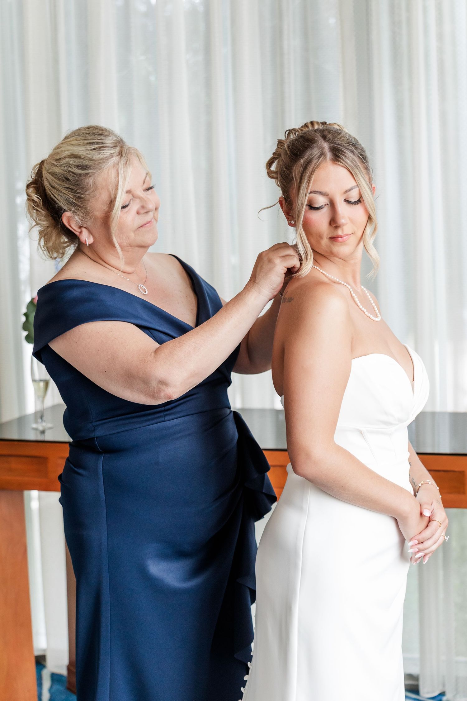 Mother in navy gown fastening pearl necklace on bride in white strapless wedding dress.