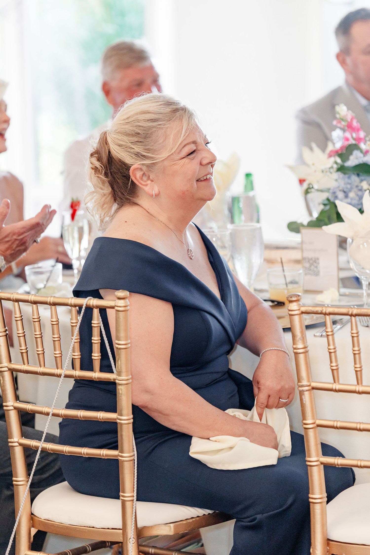 Mother of the bride smiling joyfully in navy off-shoulder gown during wedding reception speeches.