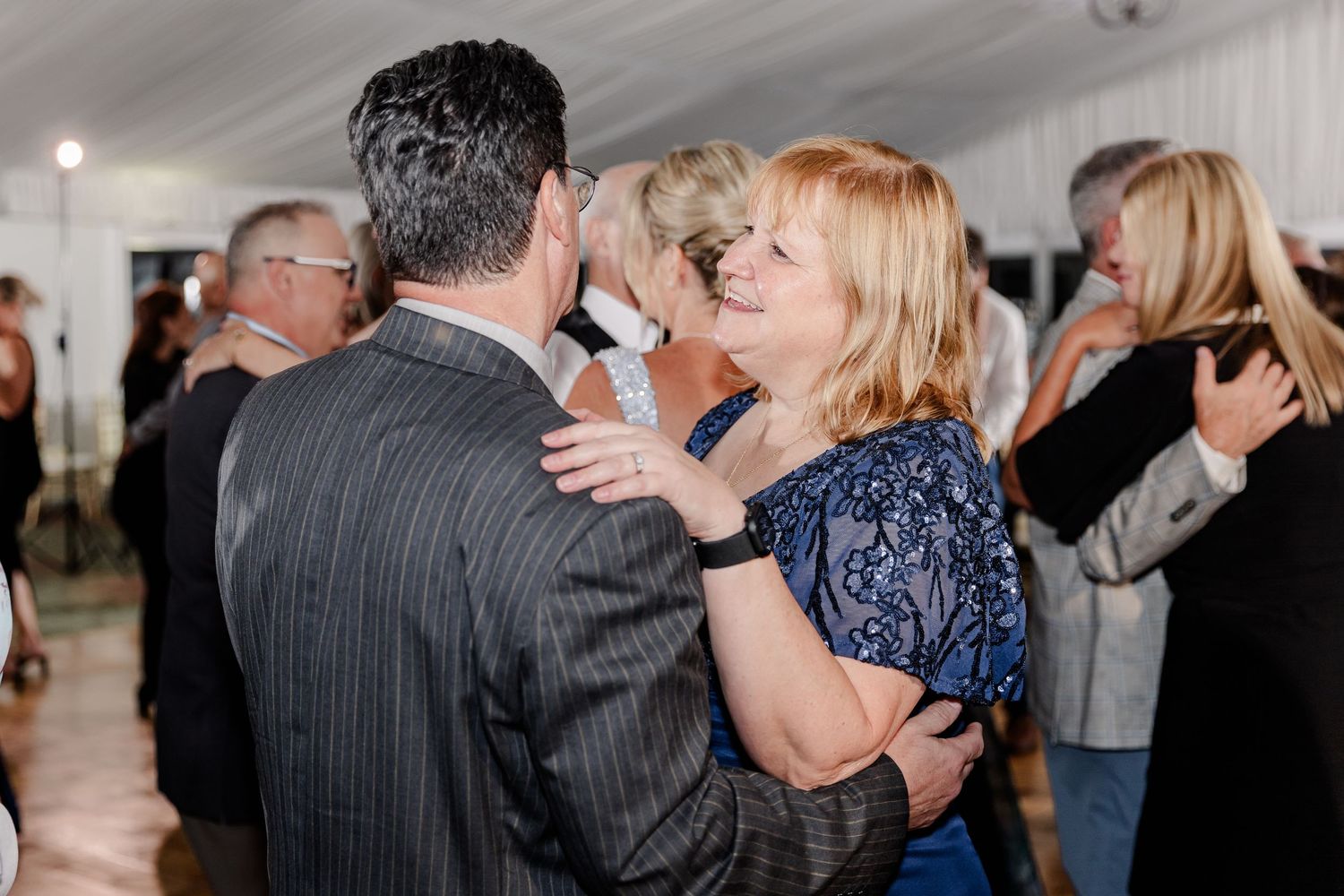 Couple slow dancing together at elegant wedding reception under white tent with other guests nearby.