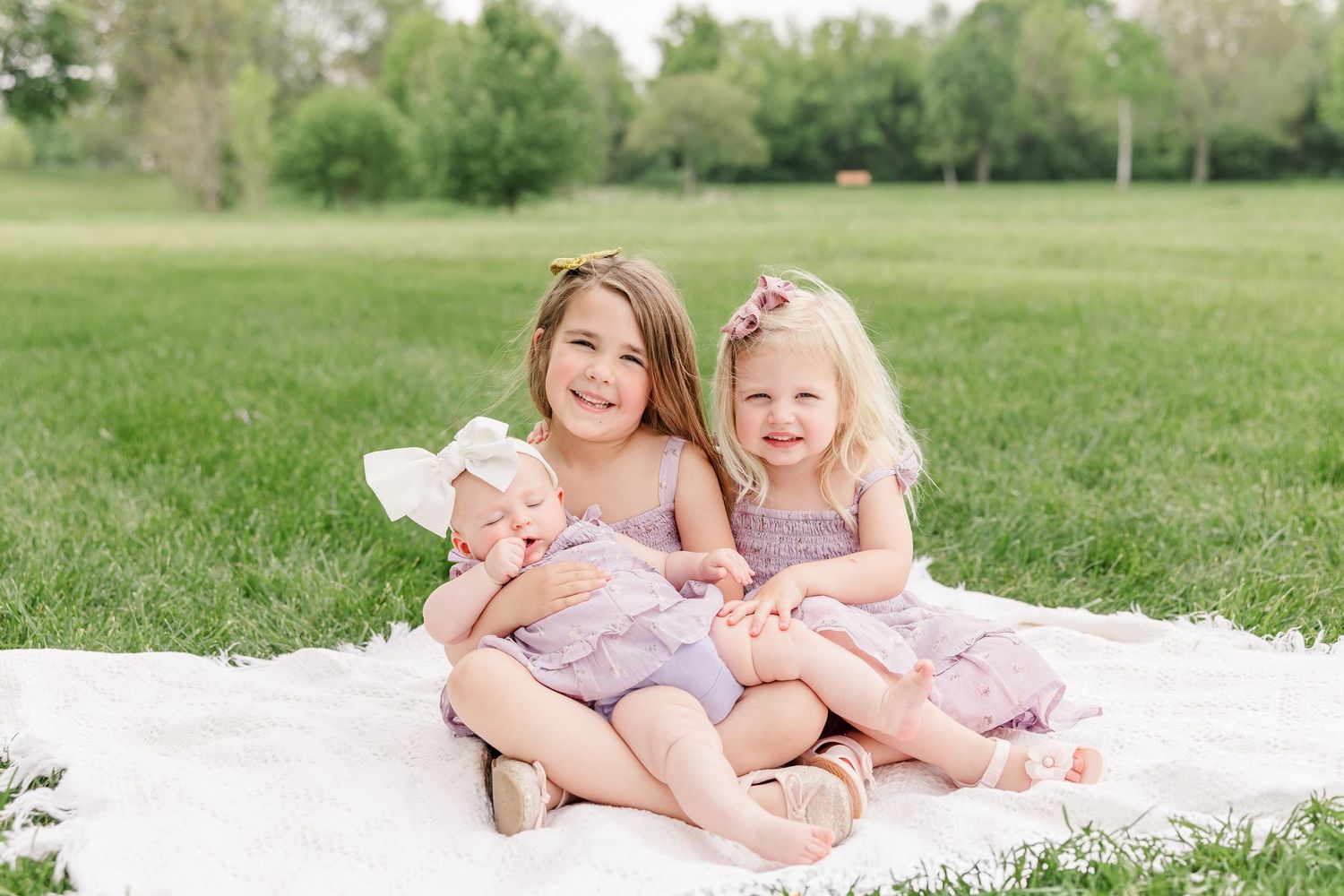Two young girls in matching lavender dresses sit on a blanket in a green park, holding a baby in a white bow.