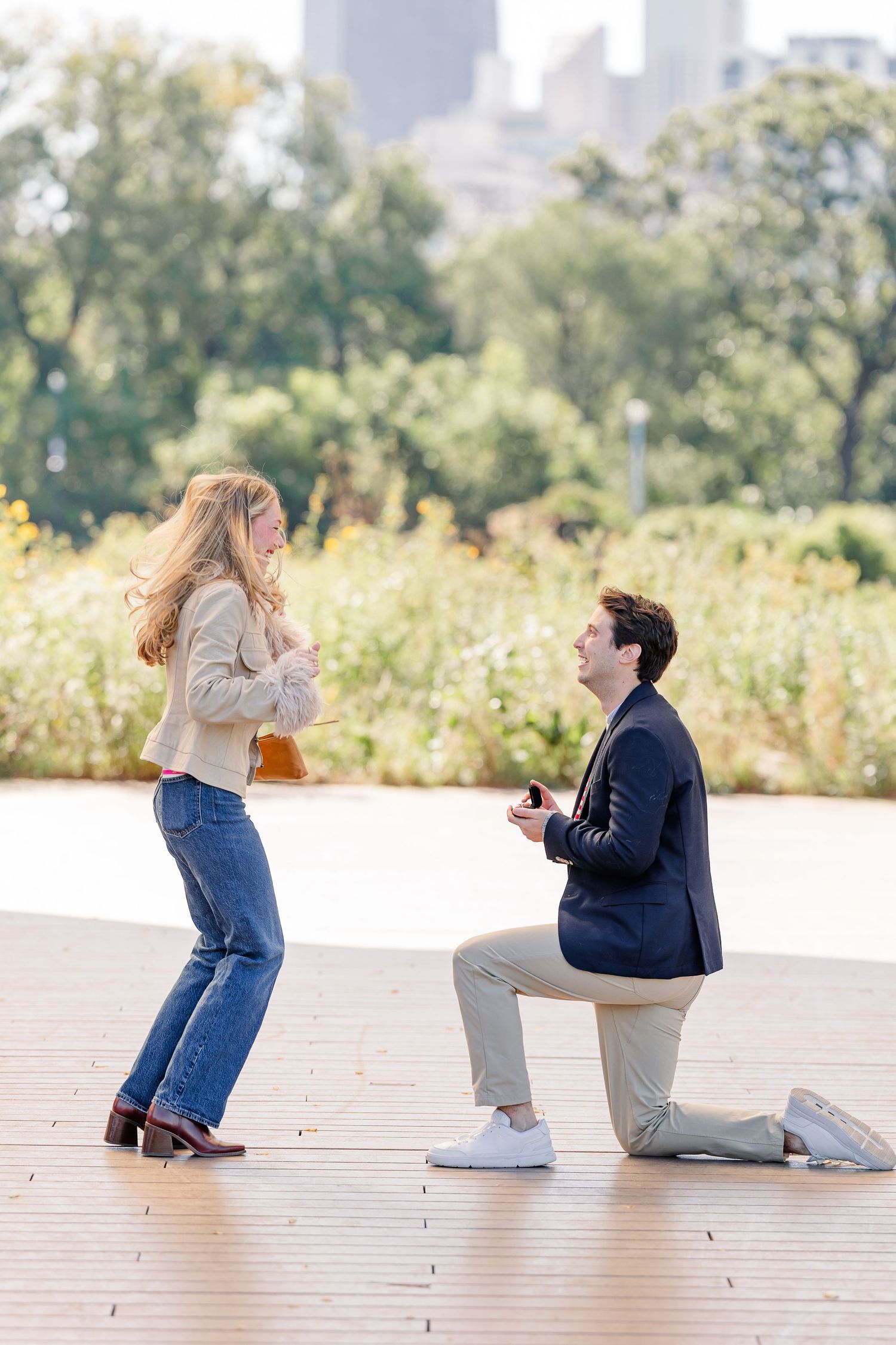 Man kneeling on one knee proposing to woman outdoors on wooden deck with lush greenery and city skyline behind.
