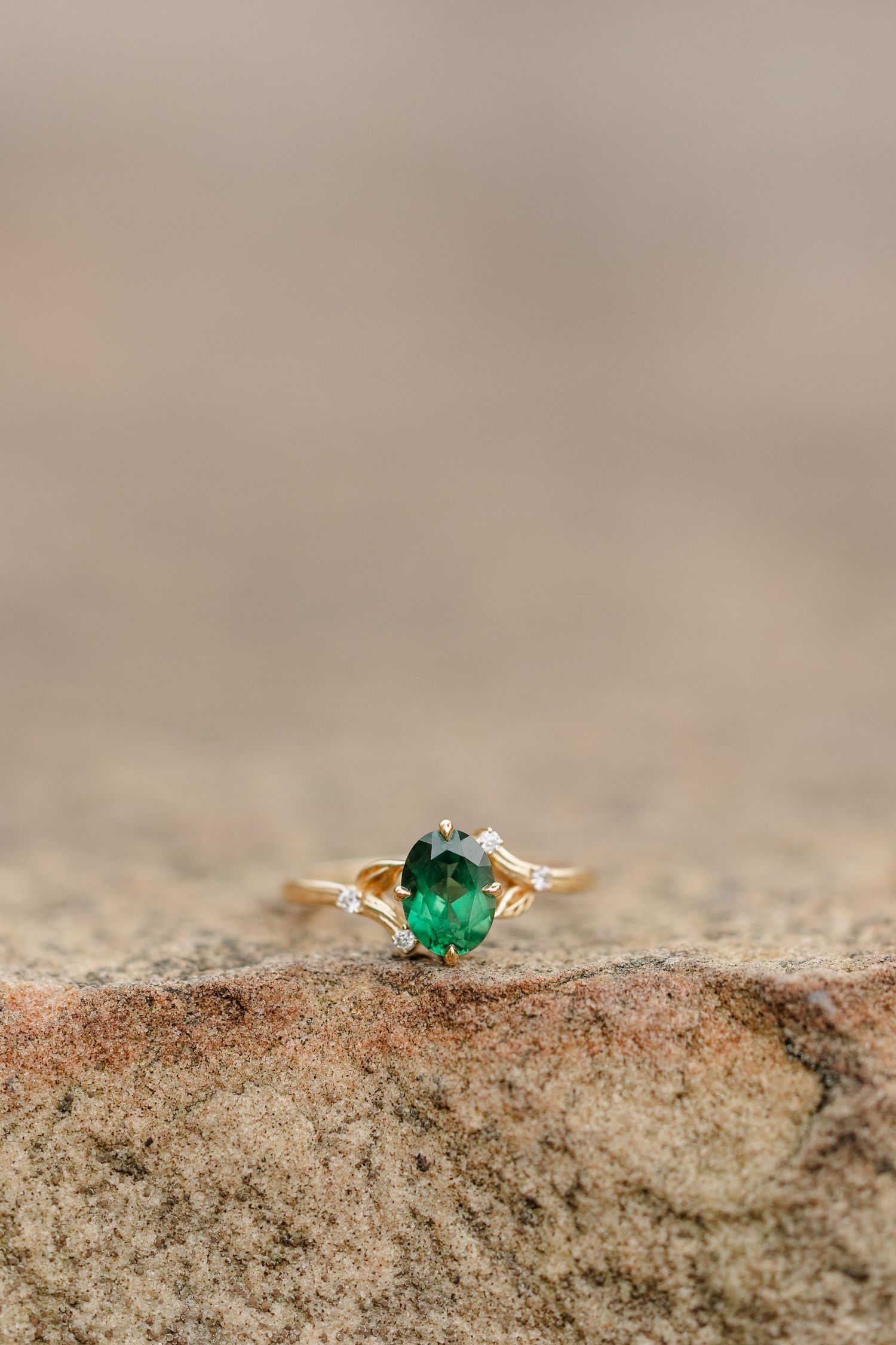 Gold engagement ring with an oval green emerald center stone and small diamond accents resting on a textured sandstone surface.