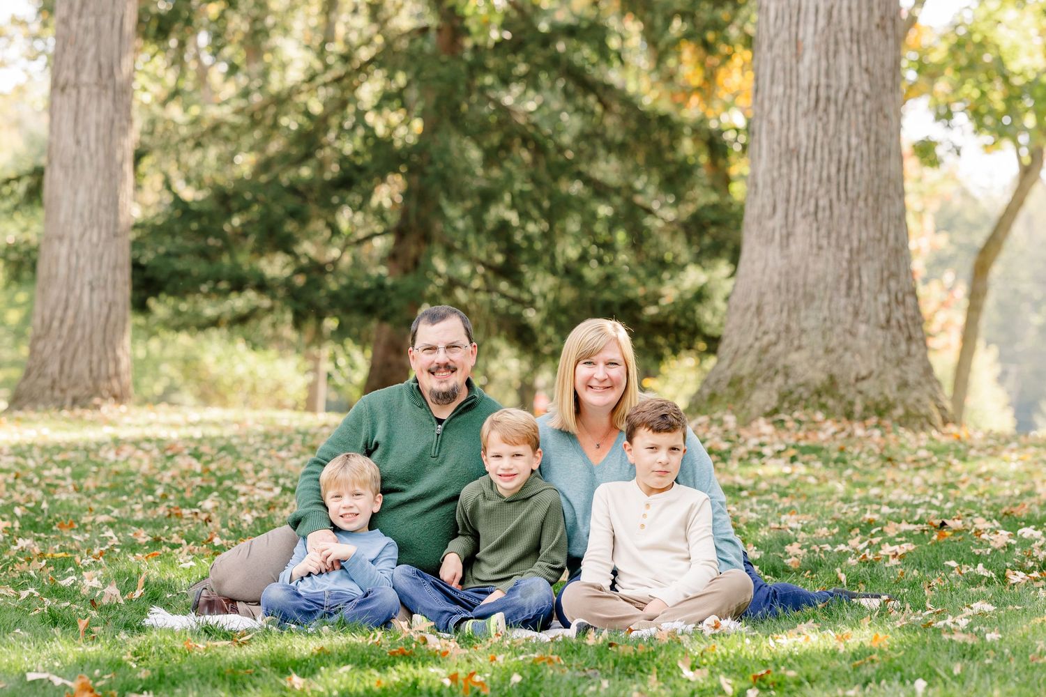 Family of five smiling outdoors on grass, two adults and three young boys seated together between large trees in a park.