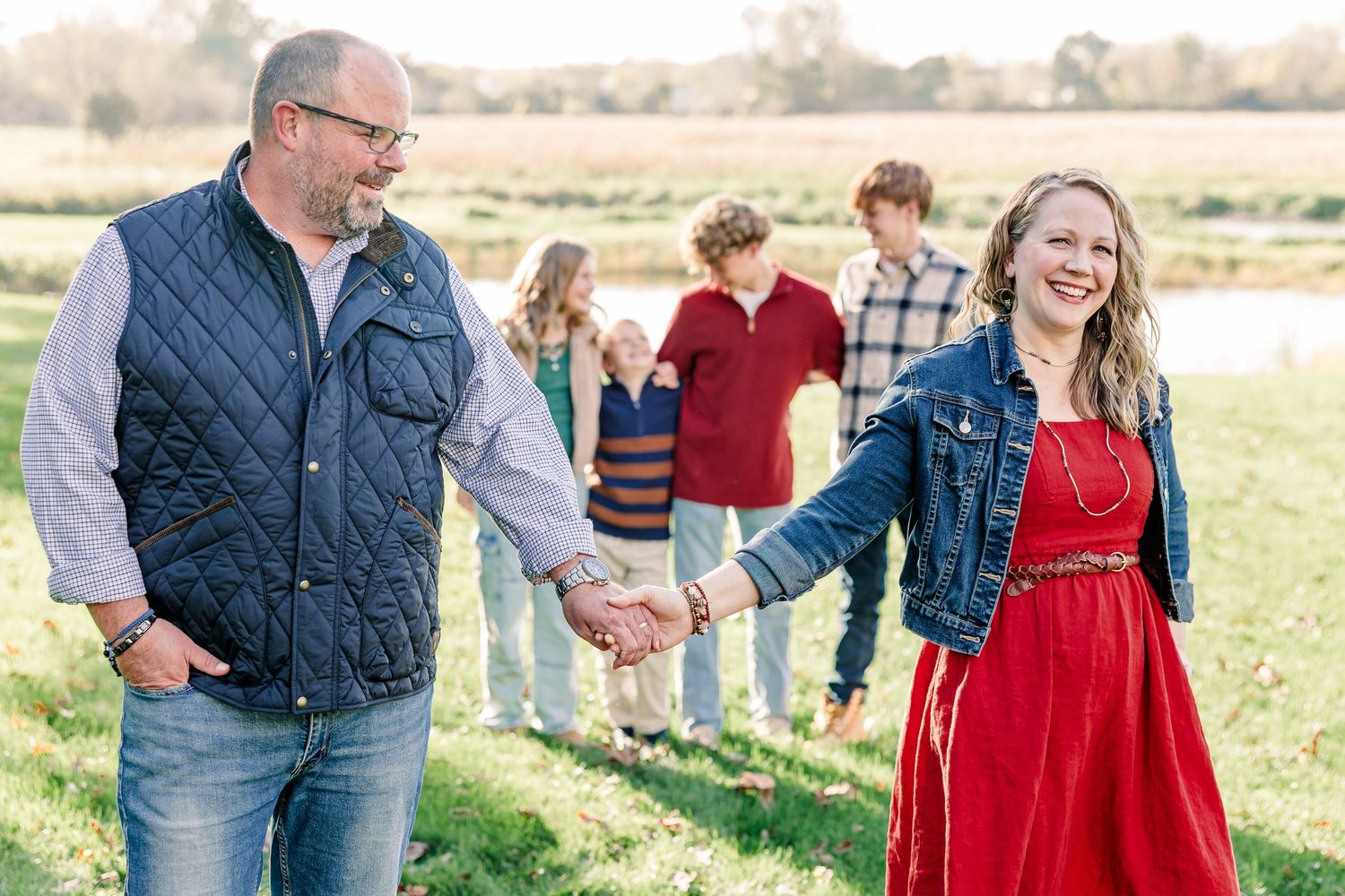 A smiling woman in a red dress and denim jacket holds hands with a bearded man in a vest as their family walks together in a grassy field.