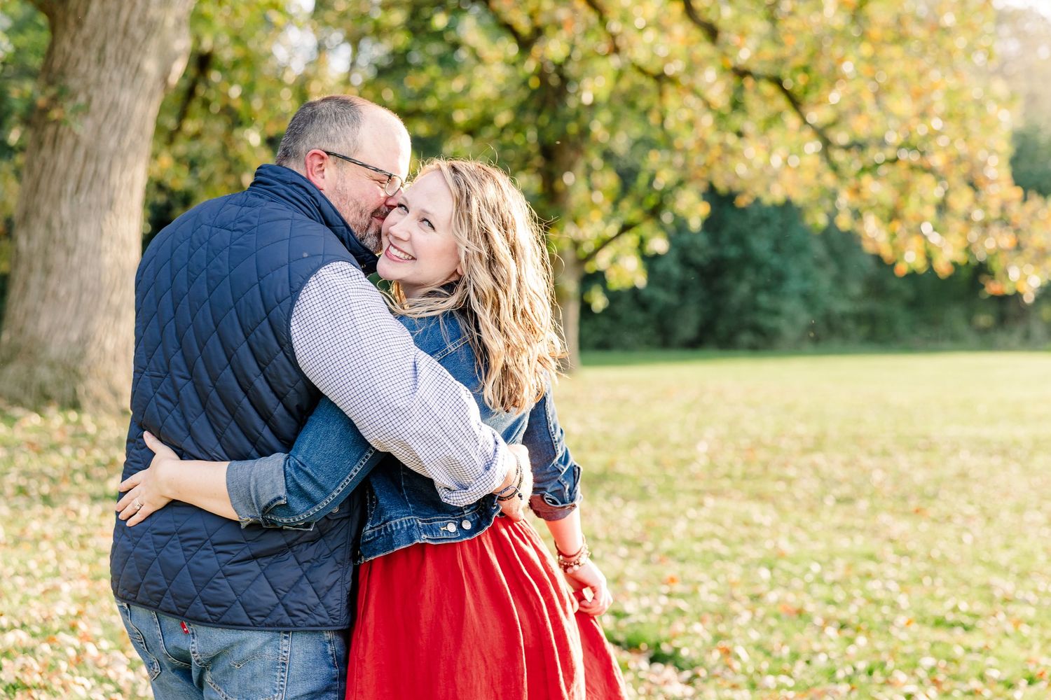 A smiling couple shares an affectionate embrace outdoors, the woman in a red skirt and denim jacket, surrounded by golden autumn trees.