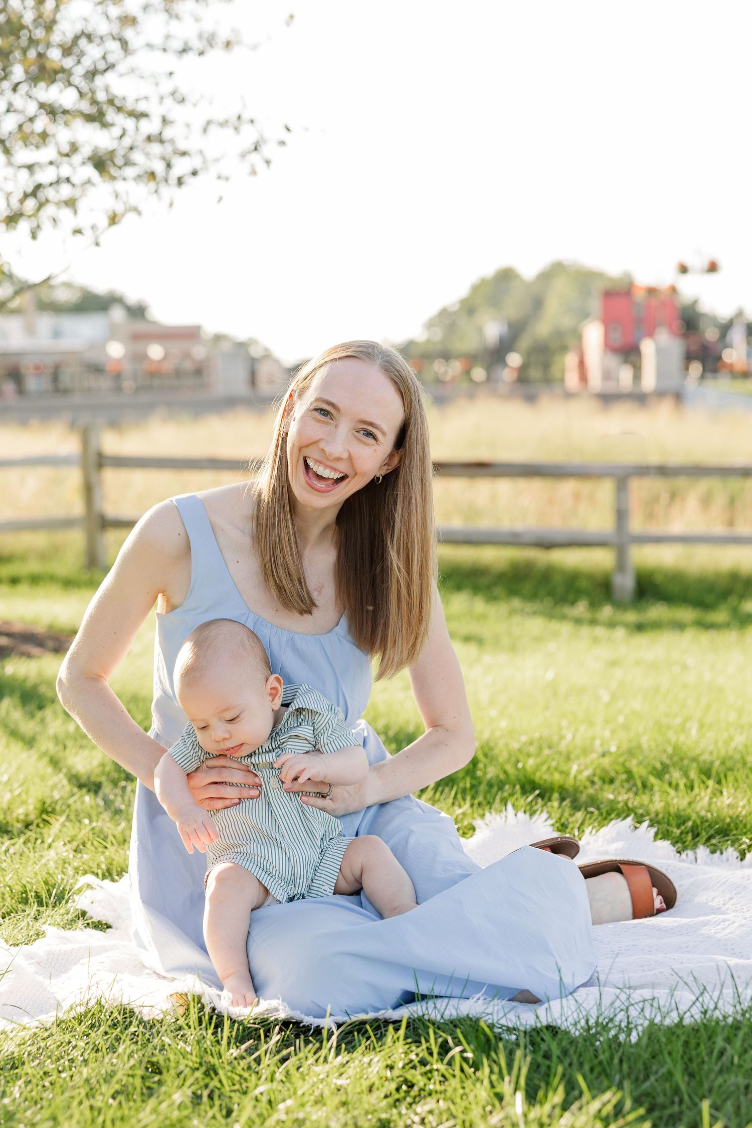 Laughing blonde mother in blue dress sitting on blanket in grass with happy baby in sunny park.