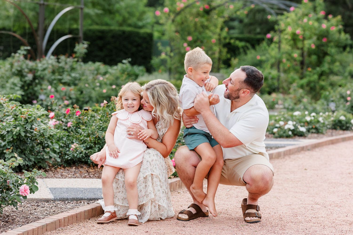Family of four poses in a rose garden, parents smiling with two young toddlers on a brick path.