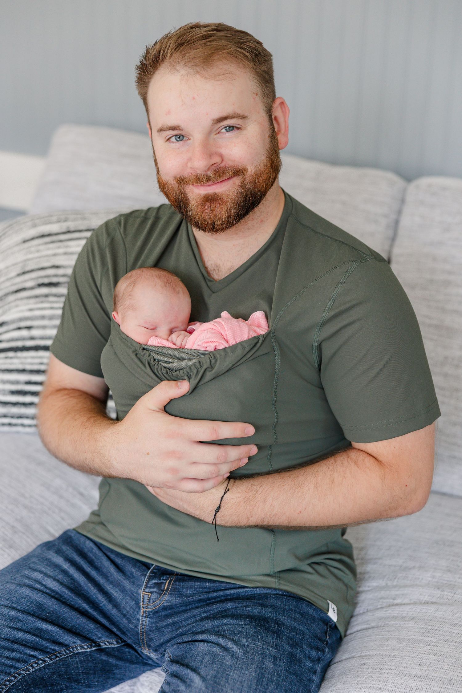Smiling red-bearded father in green t-shirt holding sleeping newborn baby in pink wrap while sitting on bed.
