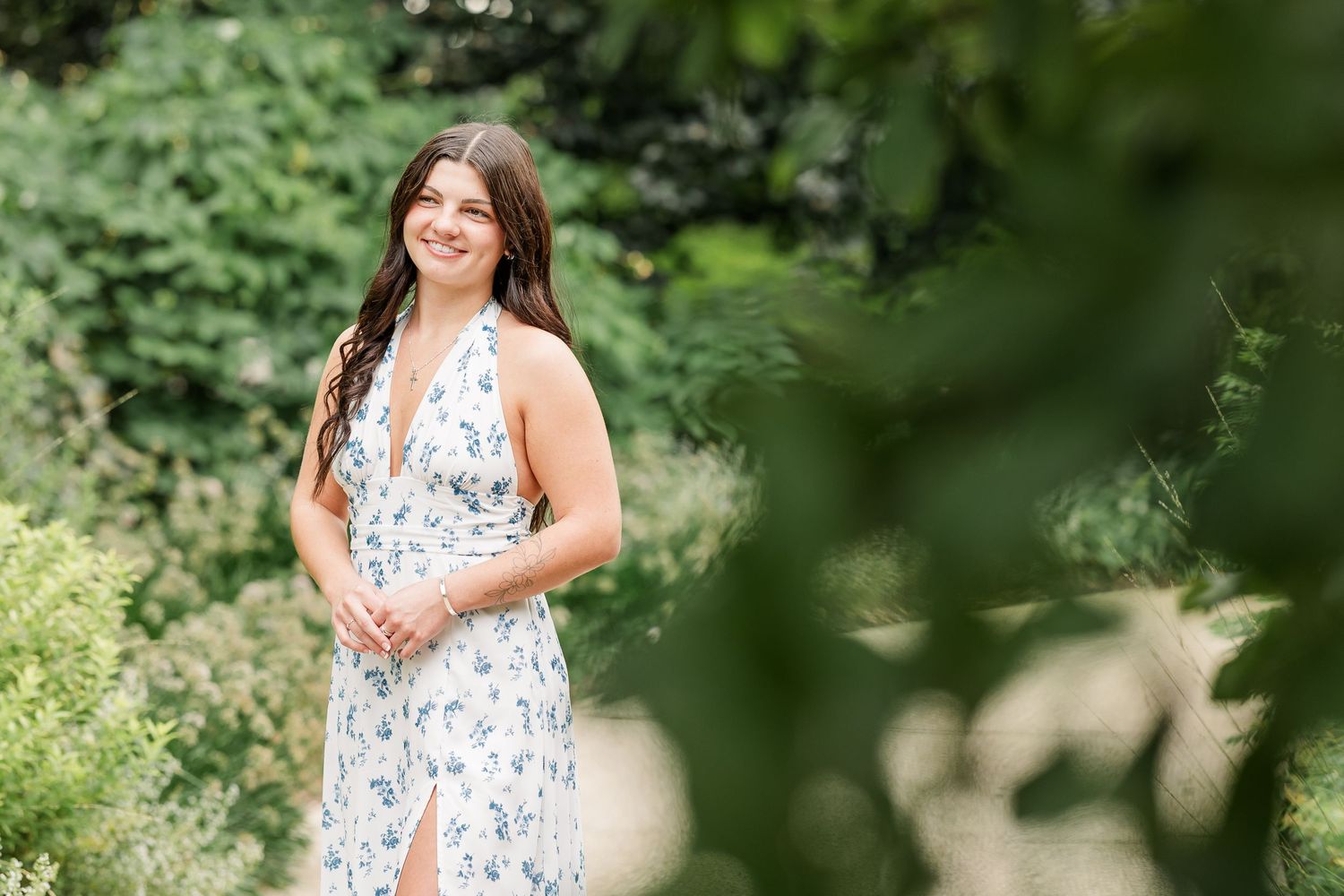 Young woman in floral halter dress smiling outdoors surrounded by lush green foliage in a garden setting.