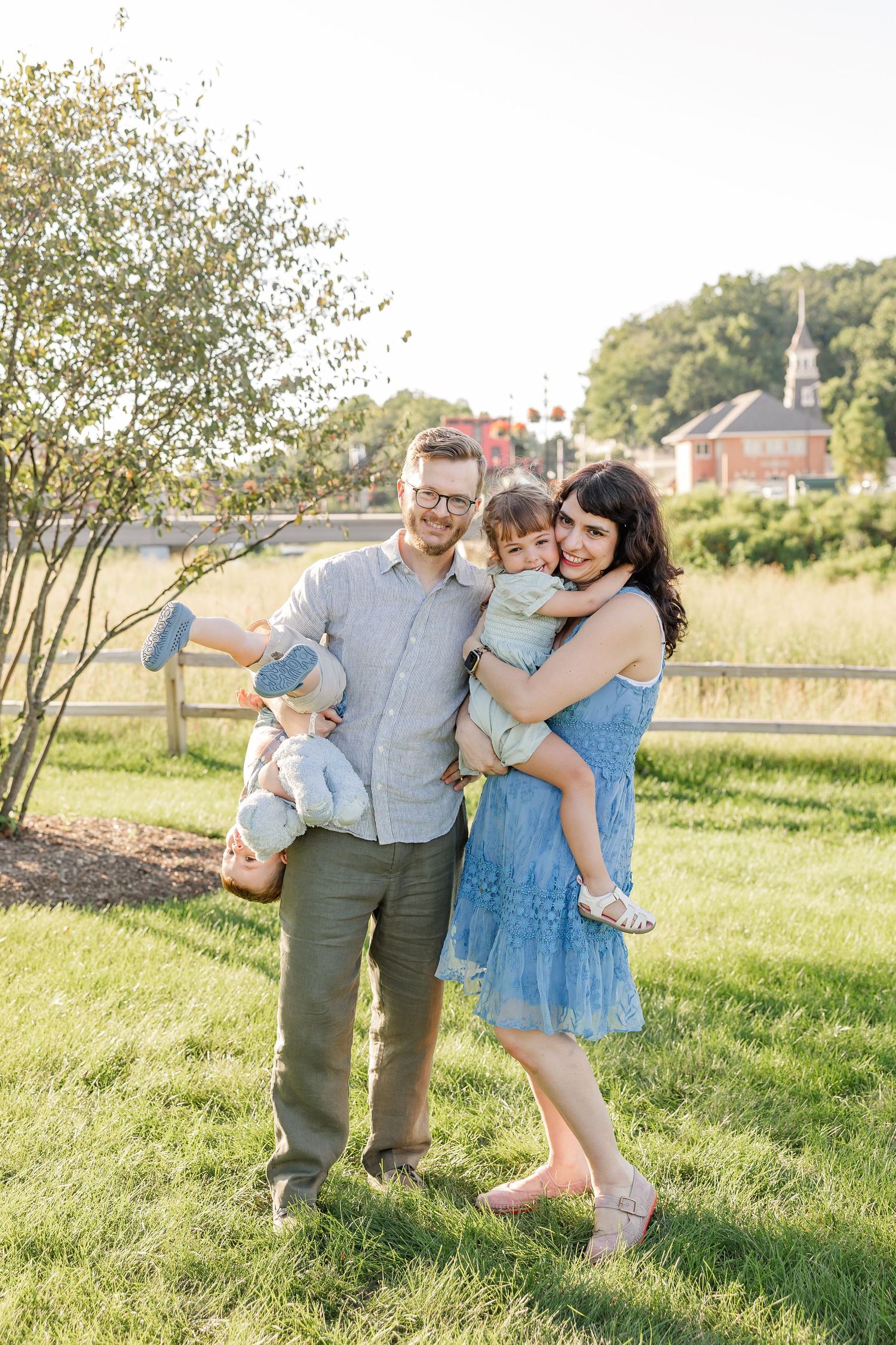 Happy family of four posing outdoors on green grass with dad holding child and mom embracing daughter.