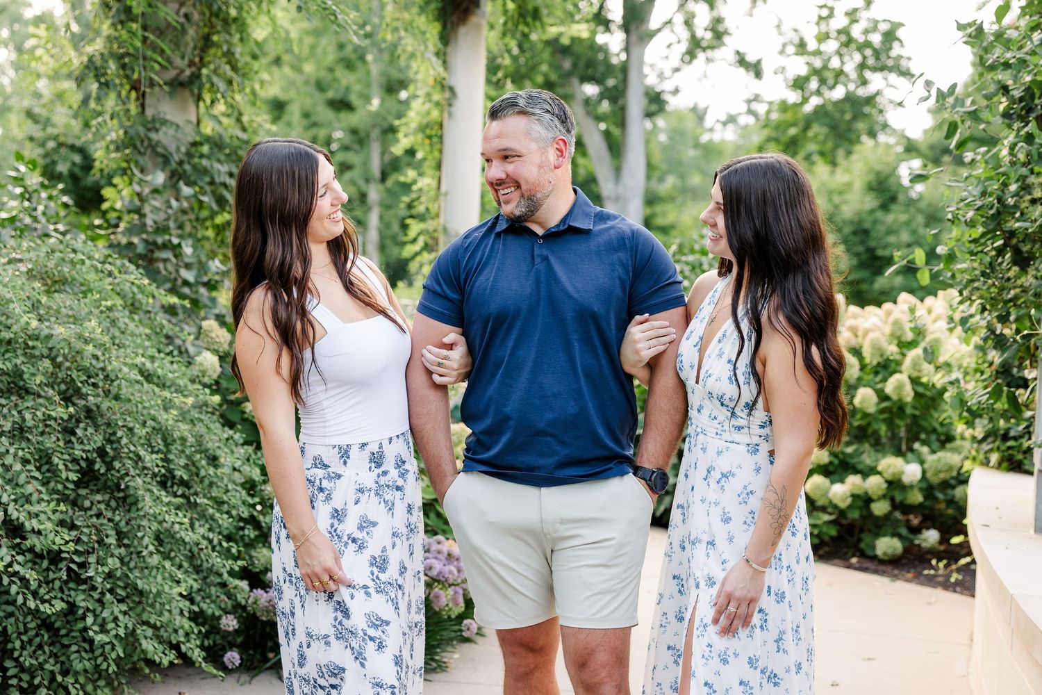 Man in navy polo with two women in floral skirts laughing together in garden setting.