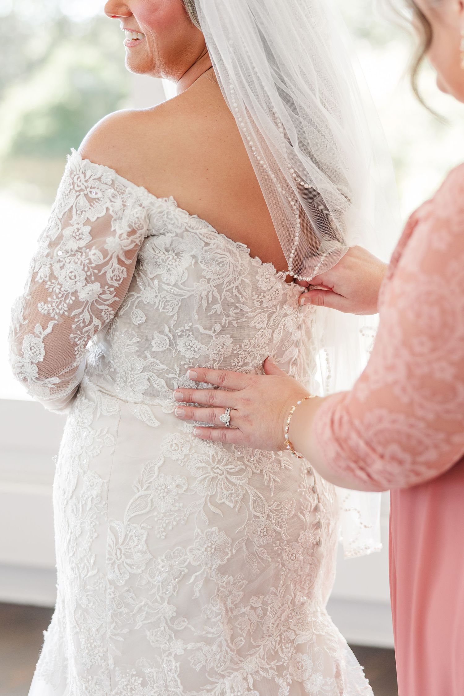 Close-up of a bridesmaid's hands fastening the covered buttons on the back of a bride's off-shoulder lace wedding dress and beaded veil.