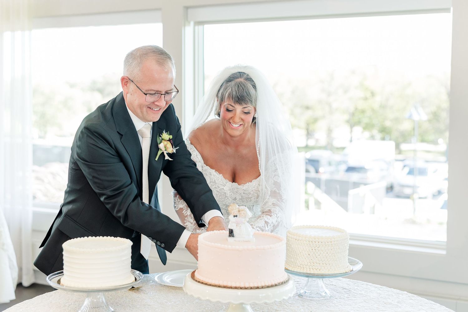 Bride in a veil and groom in a dark suit with boutonniere cut their pink wedding cake together beside a bright window.