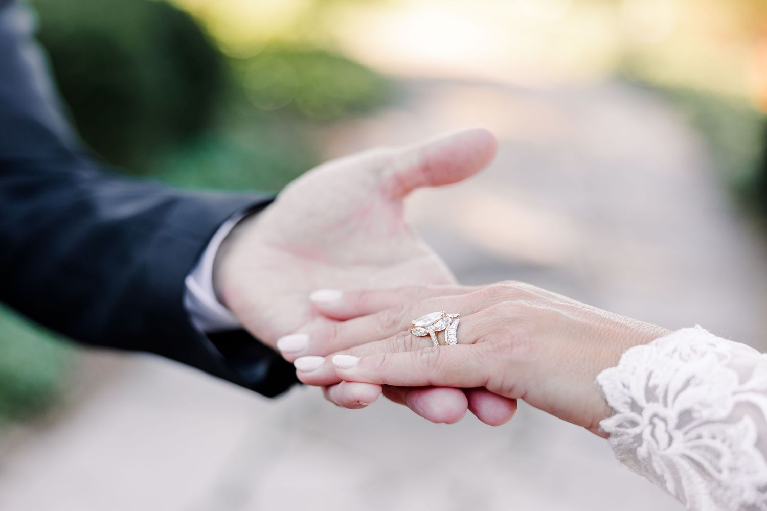 Bride in a lace sleeve placing her hand with a diamond engagement ring into the groom's outstretched hand outdoors.