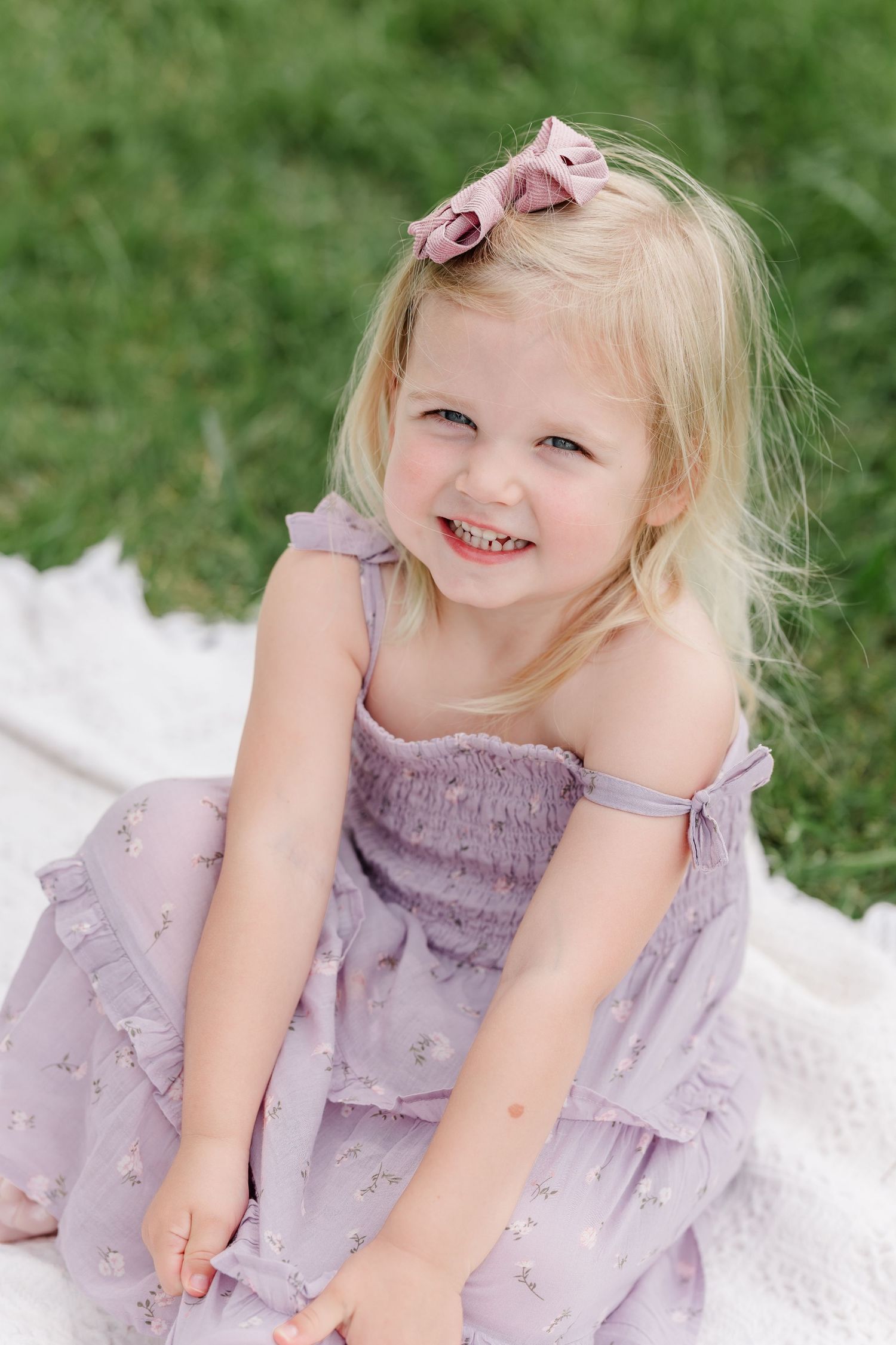 Adorable blonde toddler girl in lavender floral dress with pink bow smiling while sitting on blanket outdoors.