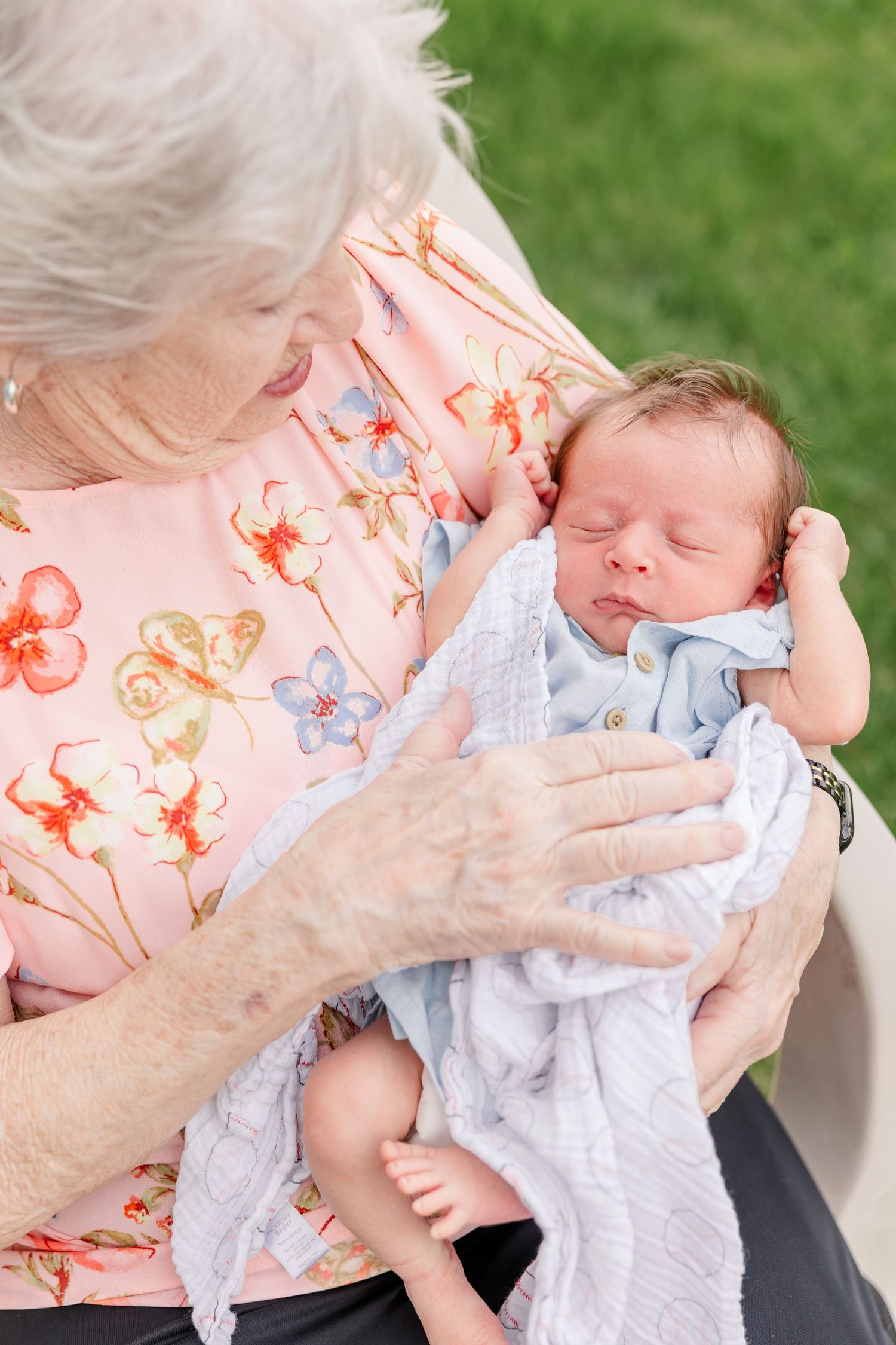 Elderly grandmother tenderly holds sleeping newborn baby wrapped in blue blanket during outdoor family photo session.