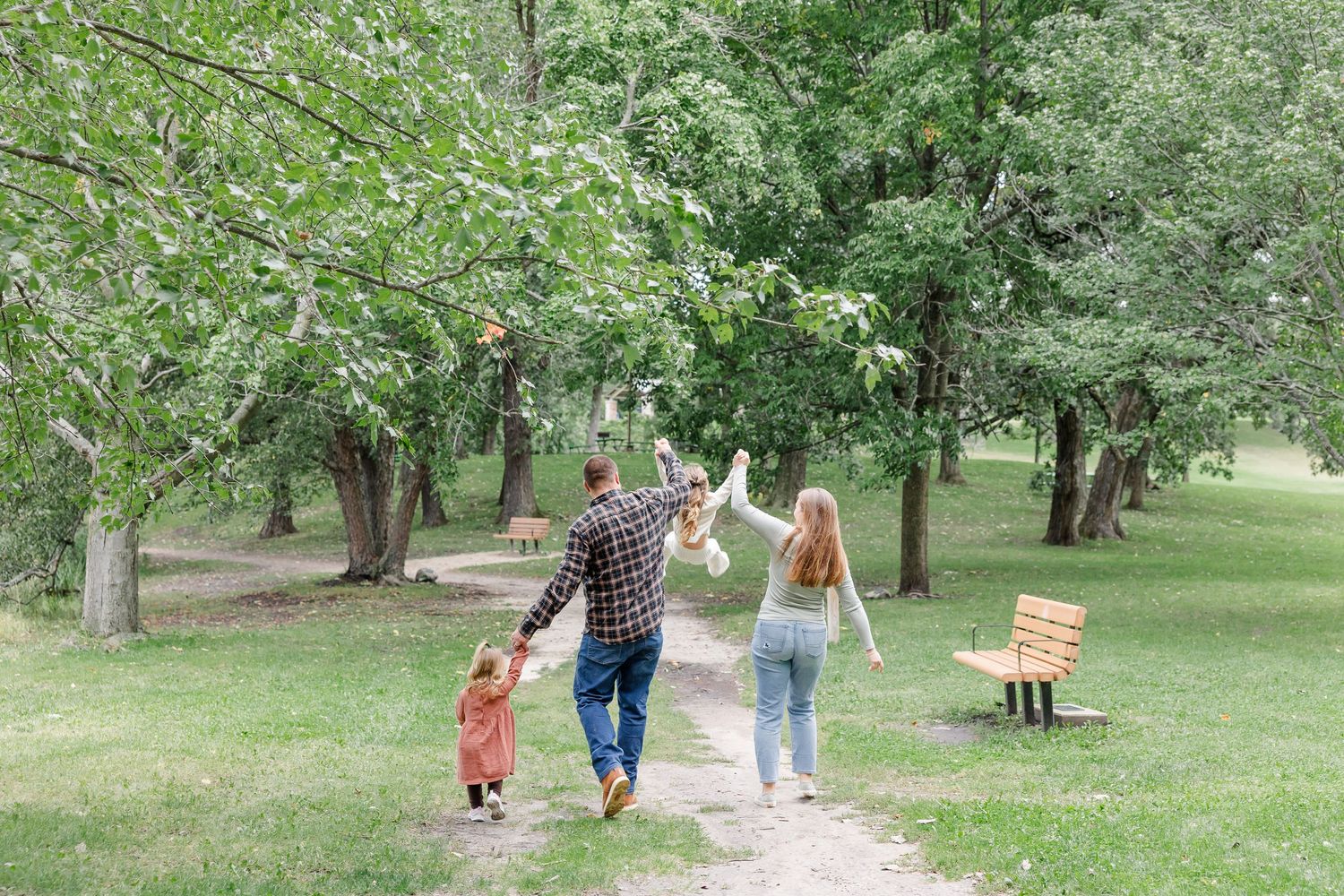 Family walking on a park path among green trees, parents holding child's hands while flying a toy airplane.