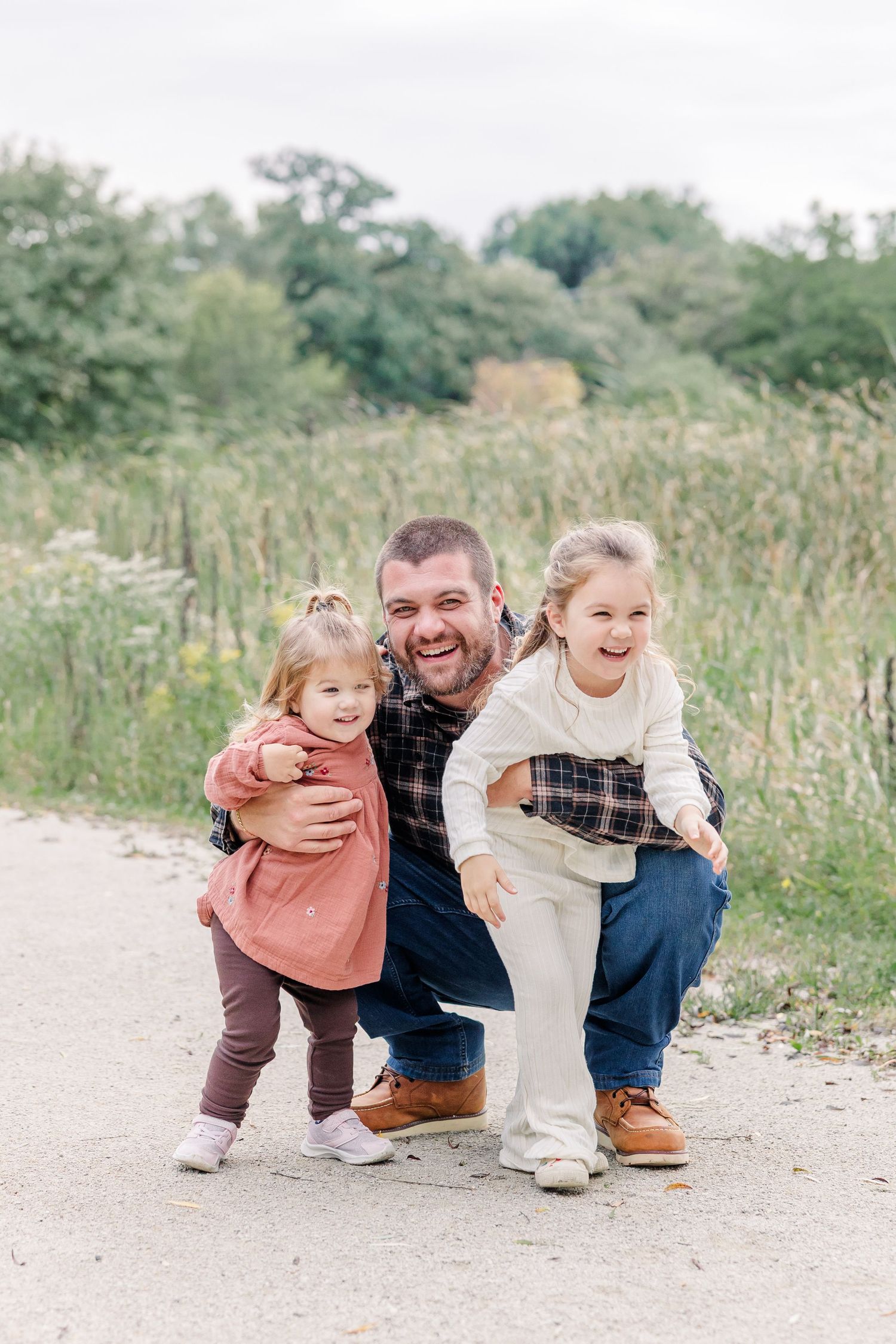 A laughing father crouches with his two young daughters on a path surrounded by lush greenery during an outdoor family photo session.