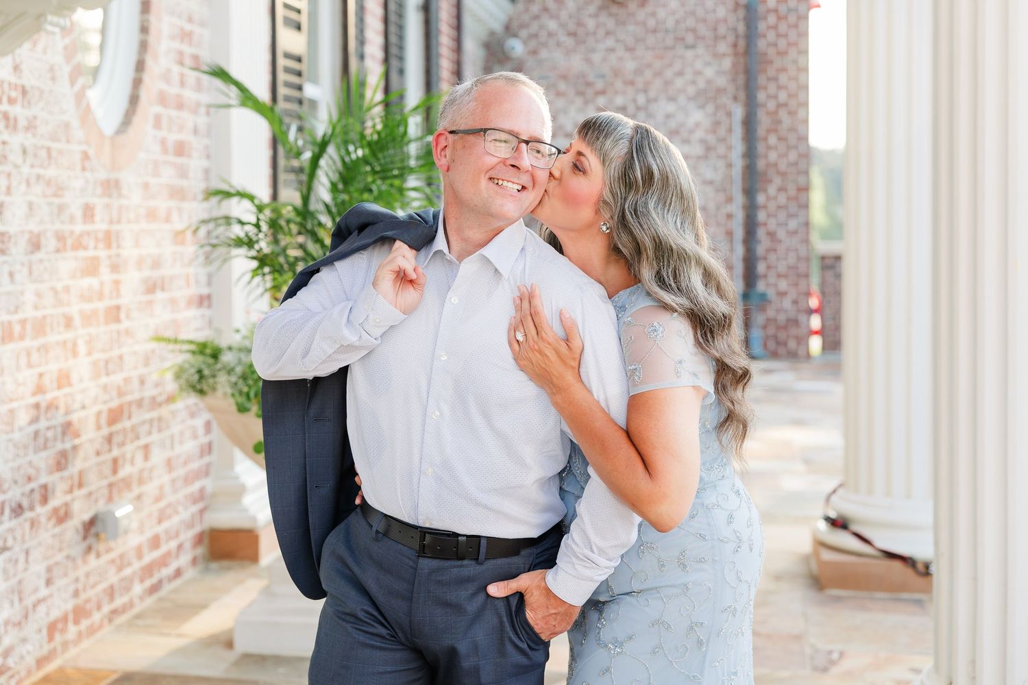 Couple sharing a romantic moment, woman kissing mans cheek outdoors near brick building with white columns.