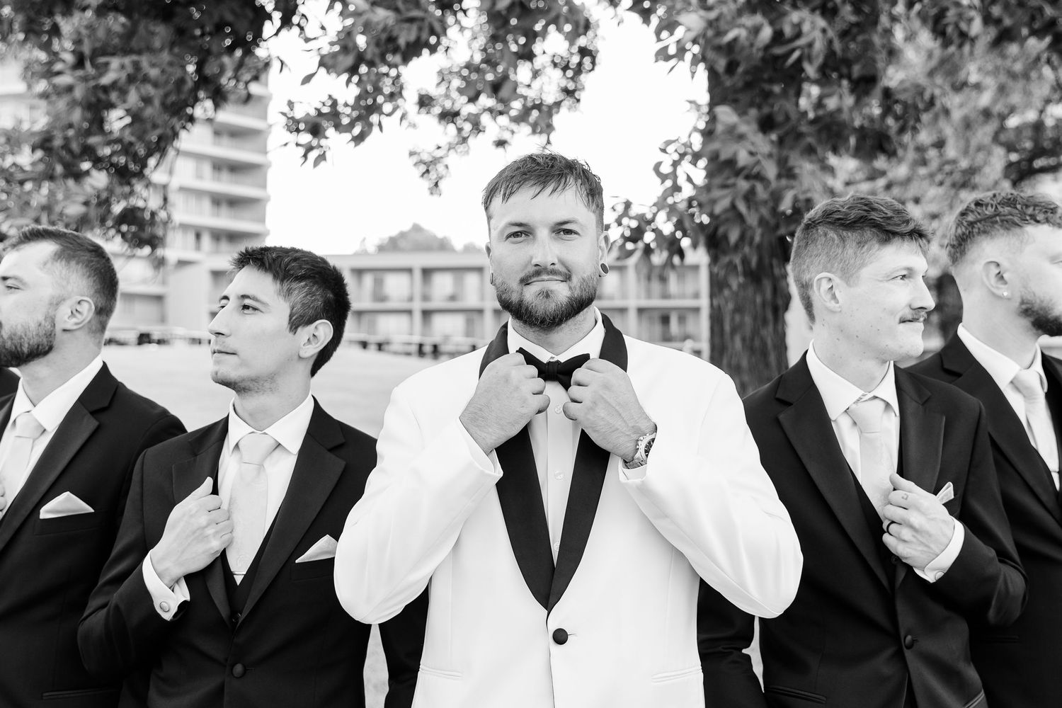 Groom in white tuxedo adjusts bow tie flanked by groomsmen in black suits, black and white photo.