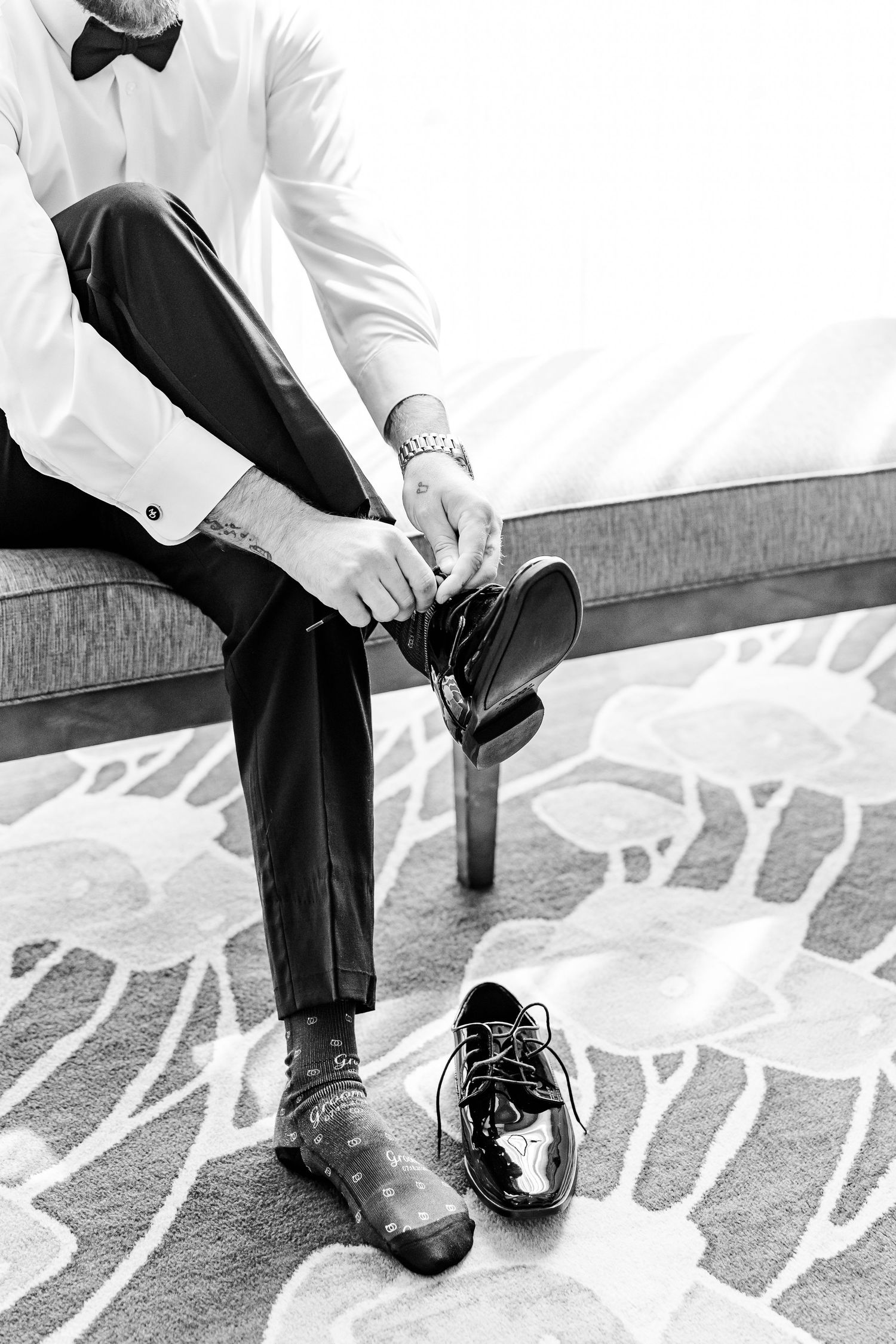 Groom in black tuxedo and white shirt putting on dress shoes while seated, black and white photo.