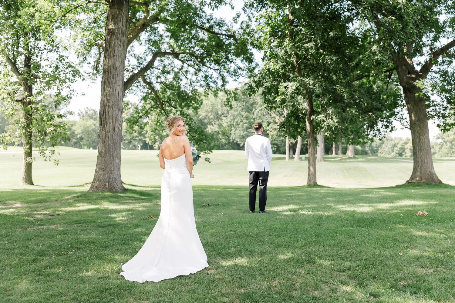 Bride in white gown and groom stand apart on lush green lawn surrounded by large trees for wedding first look.