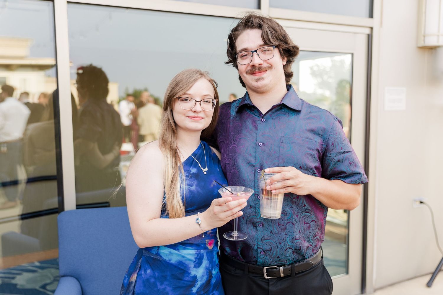 Smiling couple in formal attire holding cocktails at outdoor wedding reception celebration.