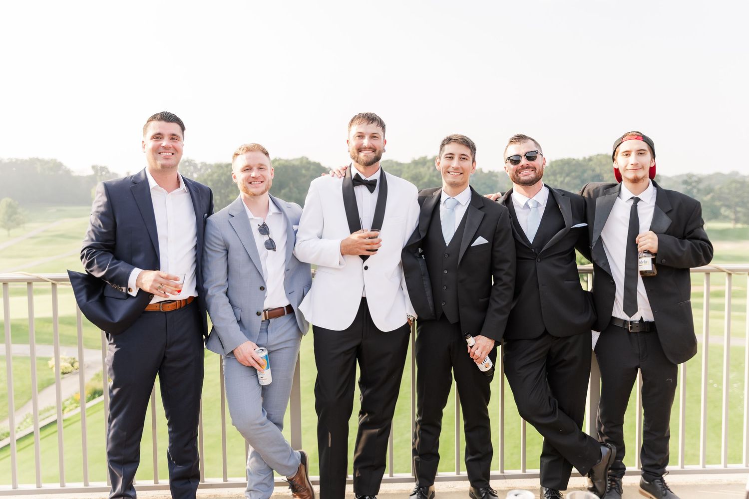 Groom in white tuxedo poses with six groomsmen on outdoor terrace overlooking golf course.