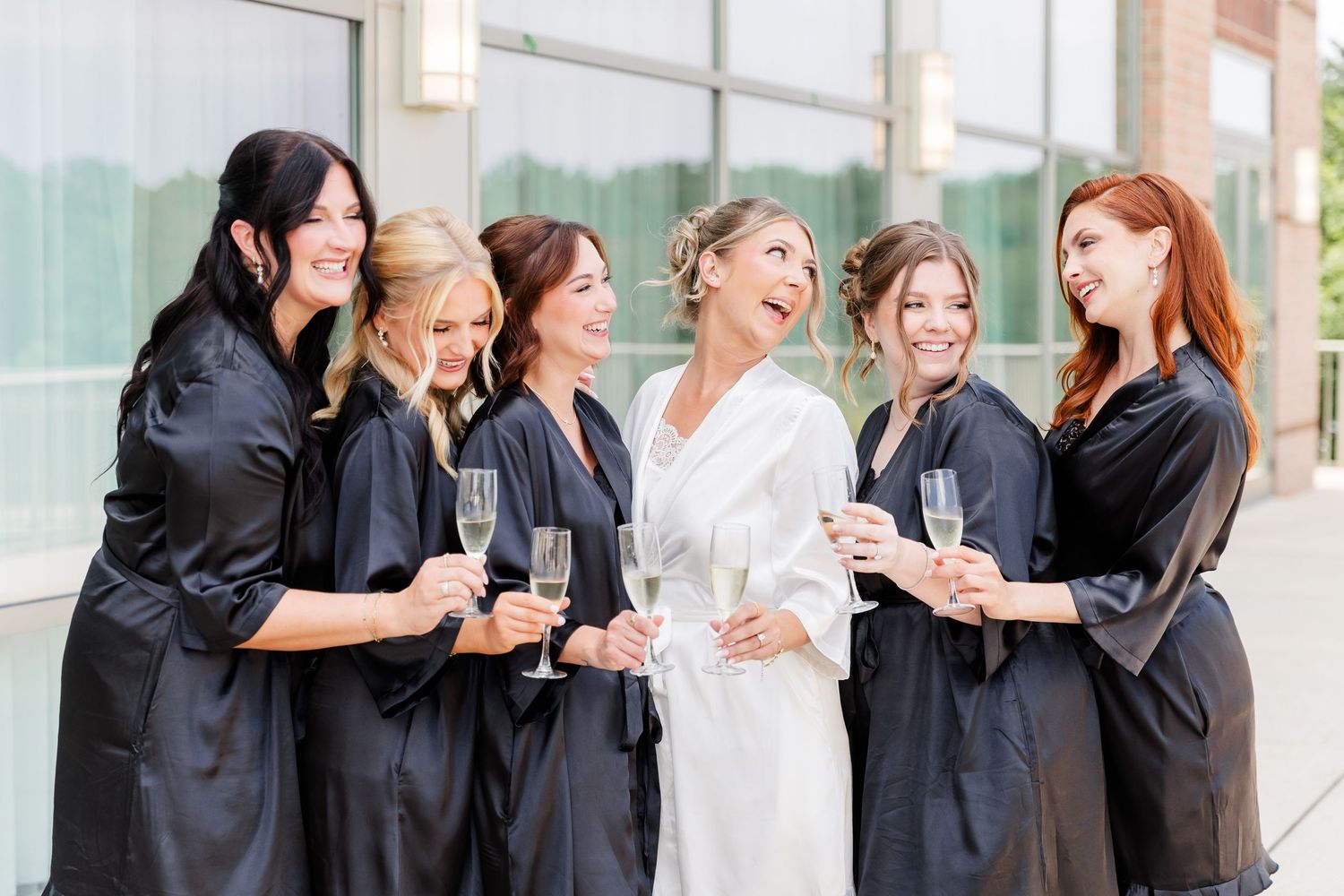 Bride and bridesmaids in black and white robes toasting with champagne glasses outdoors.