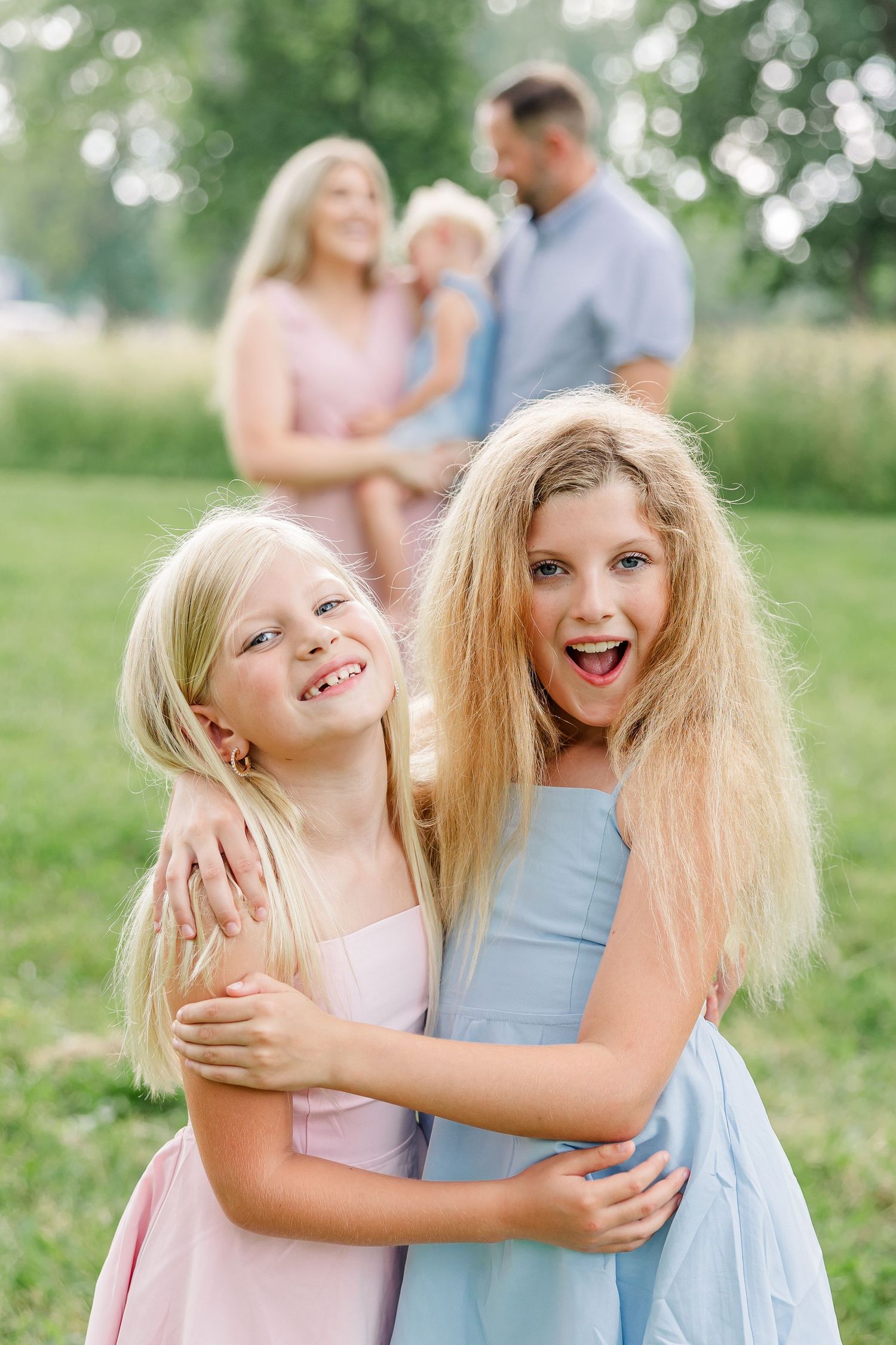Sisters hugging during a summer family session with parents in the background in Crystal Lake, IL.