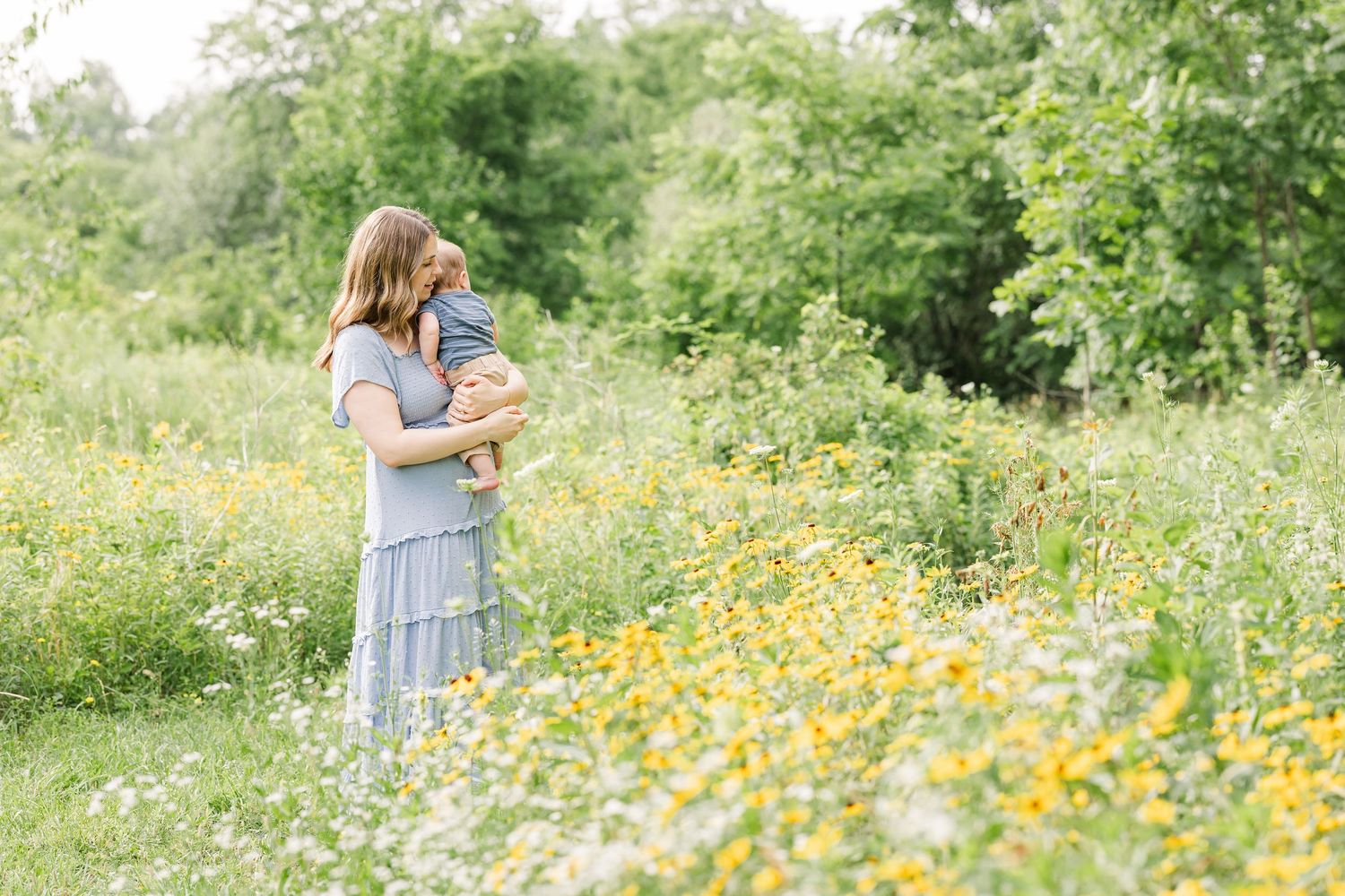 A mother holds her baby in a vibrant wildflower meadow with yellow and white blooms during an outdoor photo session.